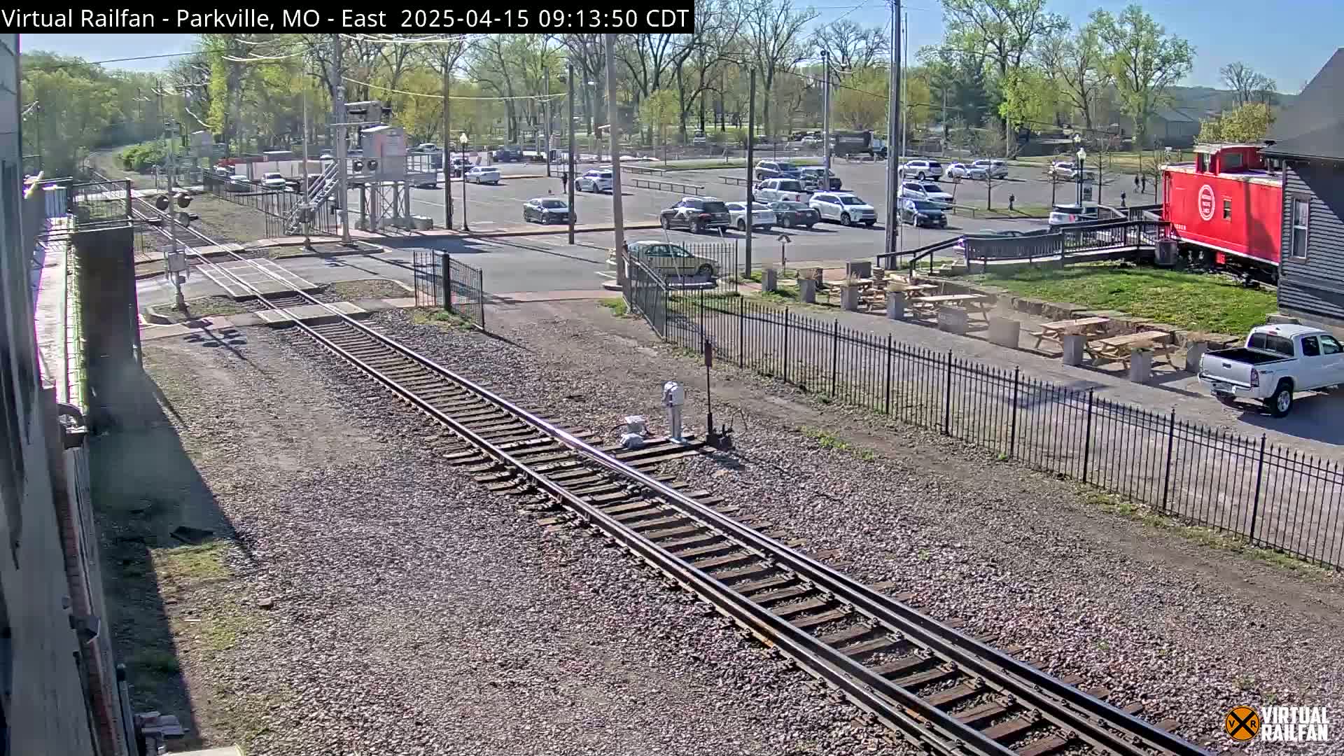 A sunny day view of railroad tracks intersecting a road with a parking lot, a red caboose, and several cars parked nearby.
