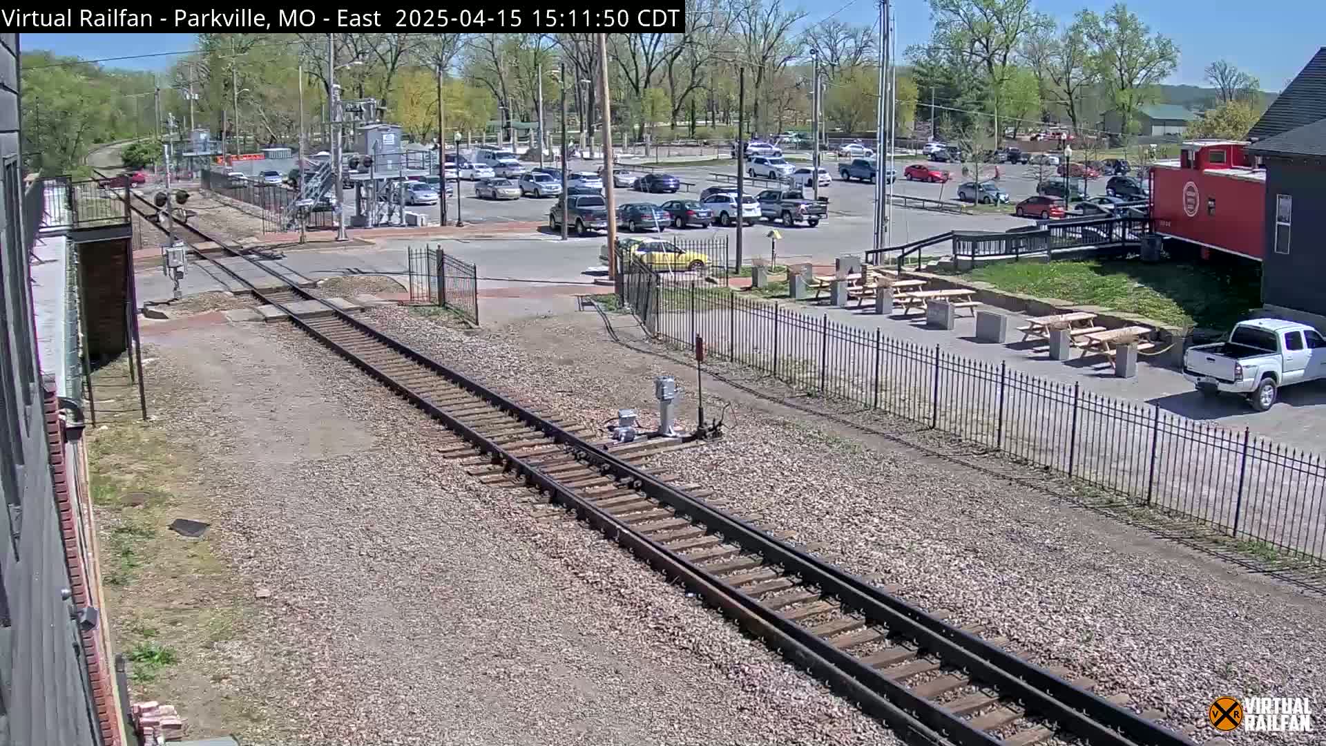A sunny day shows a railroad crossing with a parking lot full of cars, picnic tables, and a red caboose next to a building.