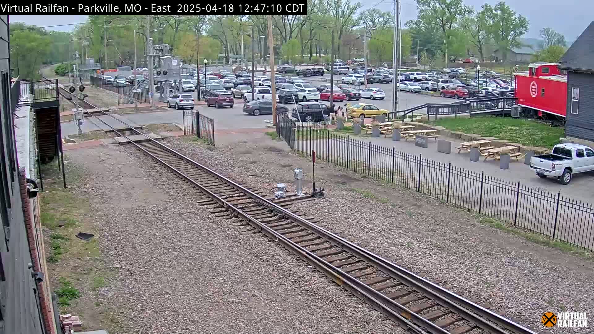 A large parking lot filled with cars is visible next to railroad tracks, a red caboose, and outdoor seating areas under a partly cloudy sky.