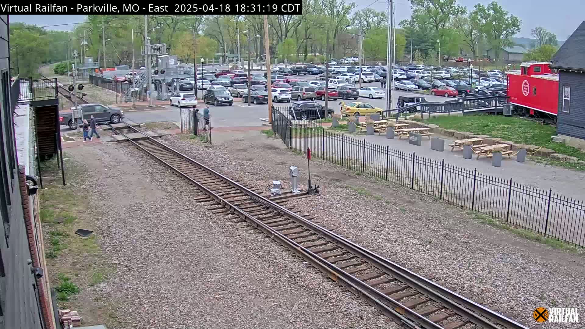 A railroad track runs through a parking lot with several cars, a red caboose, and picnic tables under a partly cloudy sky.