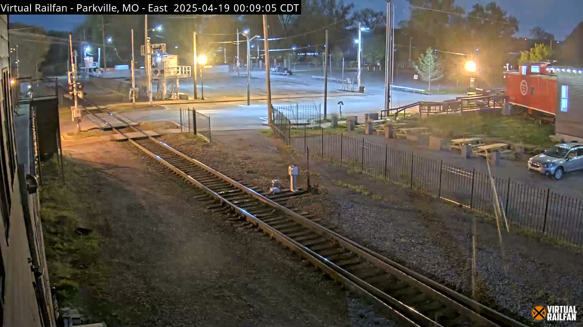 A nighttime view of railroad tracks intersecting a road near a building and outdoor seating area under partly cloudy skies.