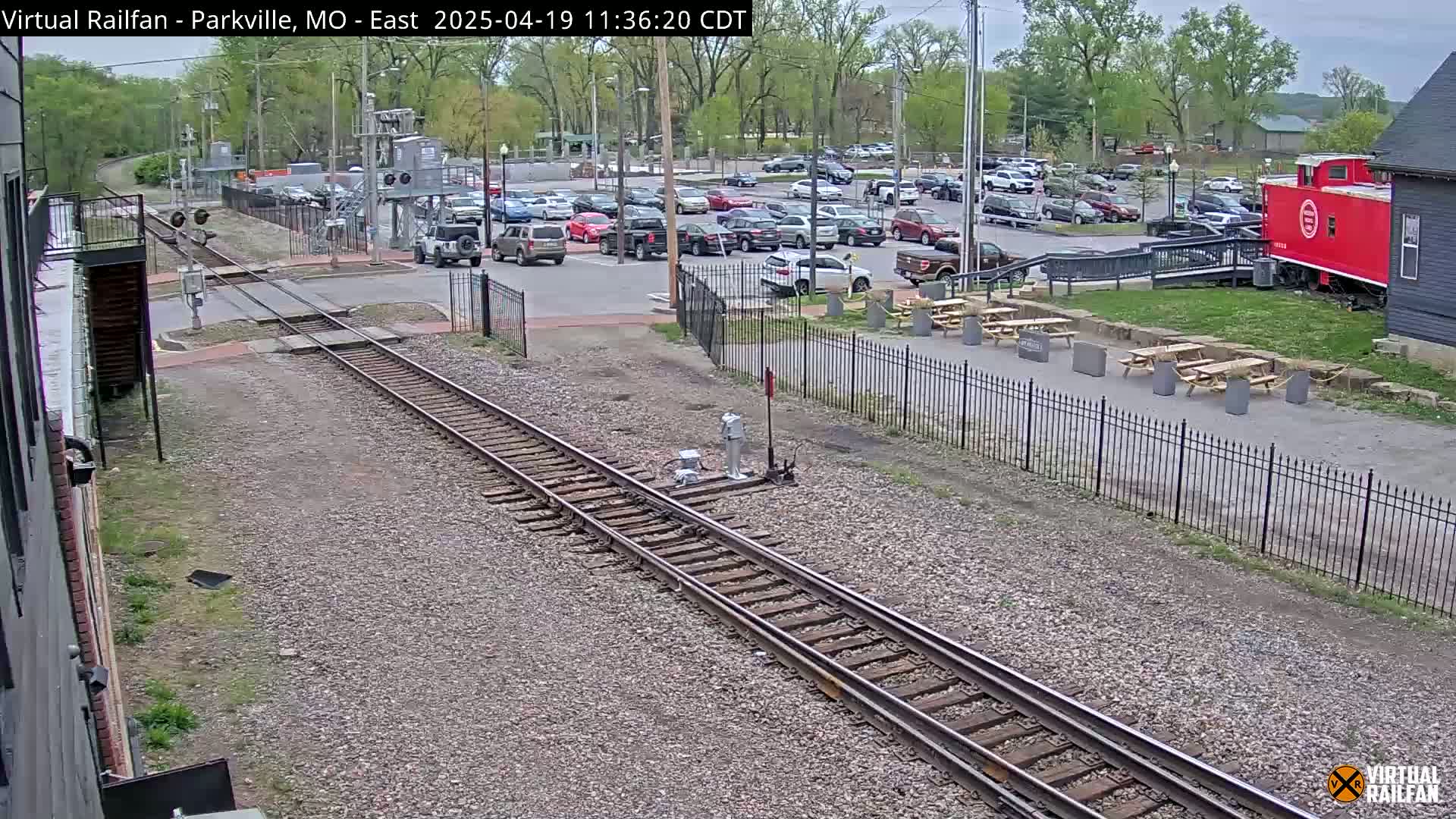 Railroad tracks extend from the foreground past a parking lot with numerous cars, a red caboose, and several picnic tables under a partly cloudy sky.
