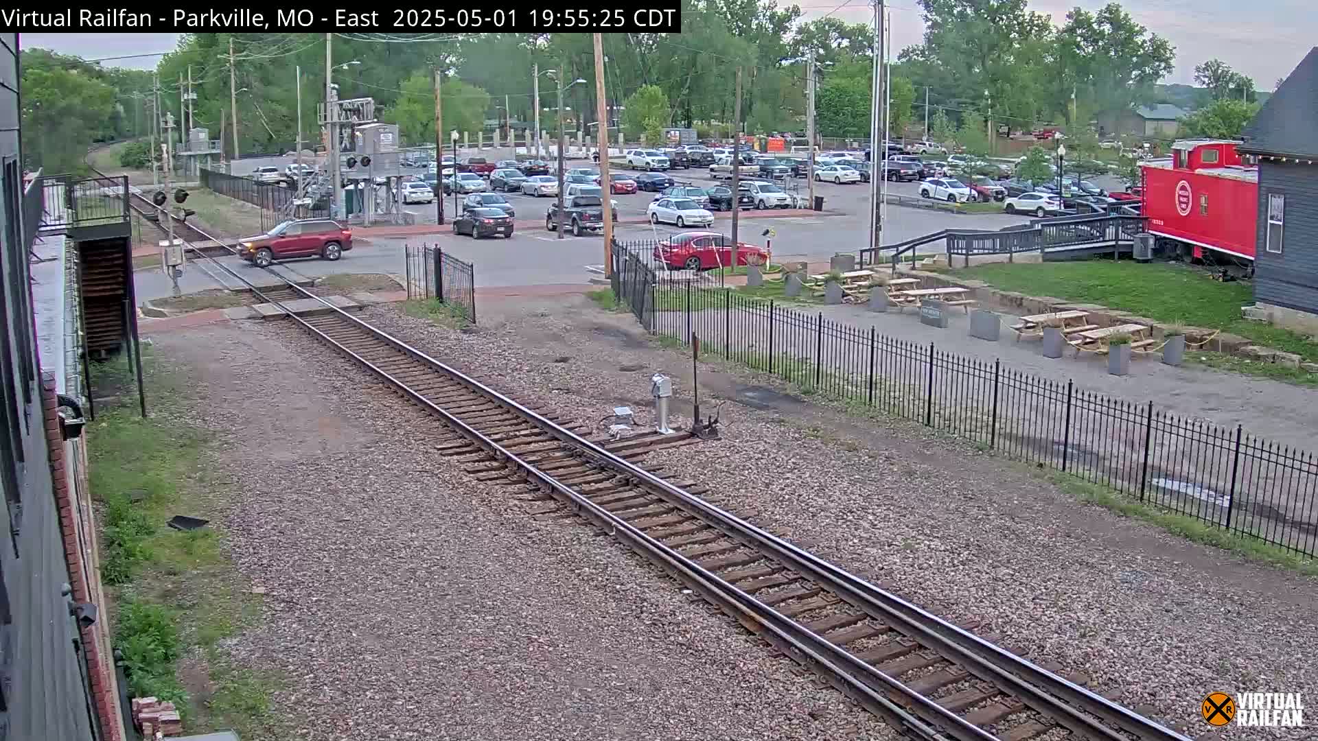 A red SUV crosses a railroad crossing near a parking lot and a red caboose under a partly cloudy sky.