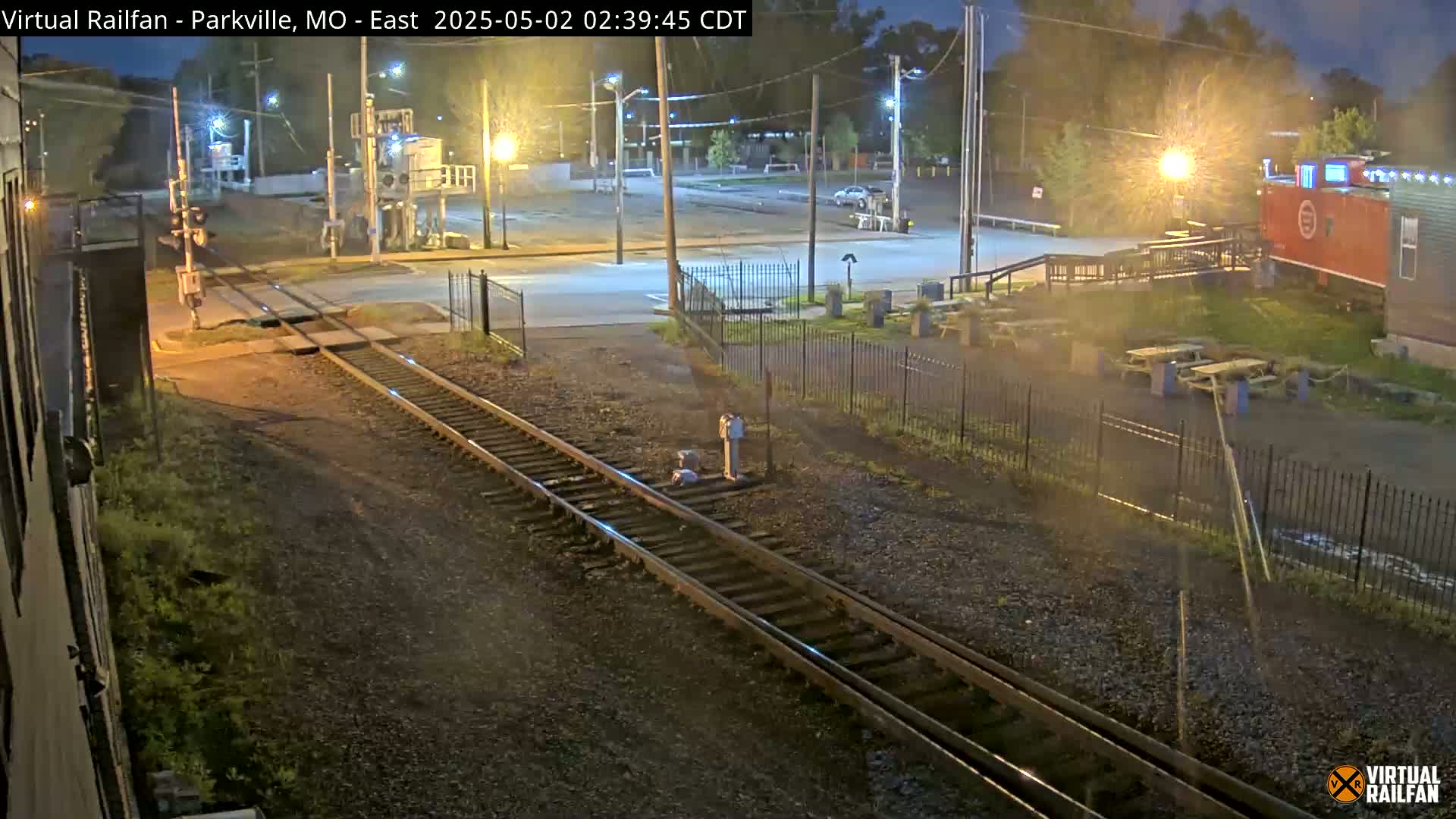 A nighttime view of railroad tracks intersecting a road, with a building and outdoor seating area visible in the background under partly cloudy skies.