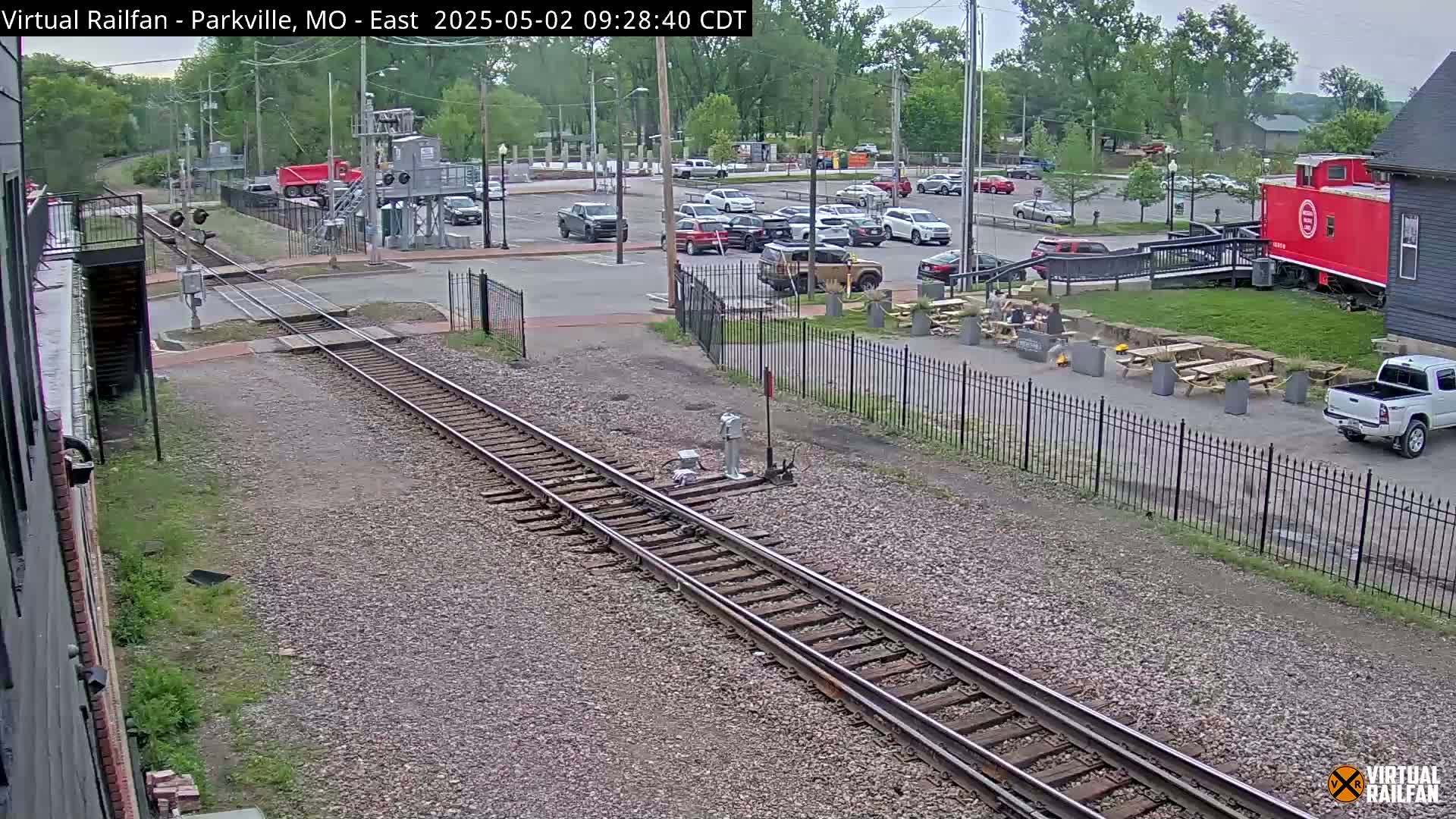 Railroad tracks run alongside a parking lot with cars and a building featuring outdoor seating under a partly cloudy sky.