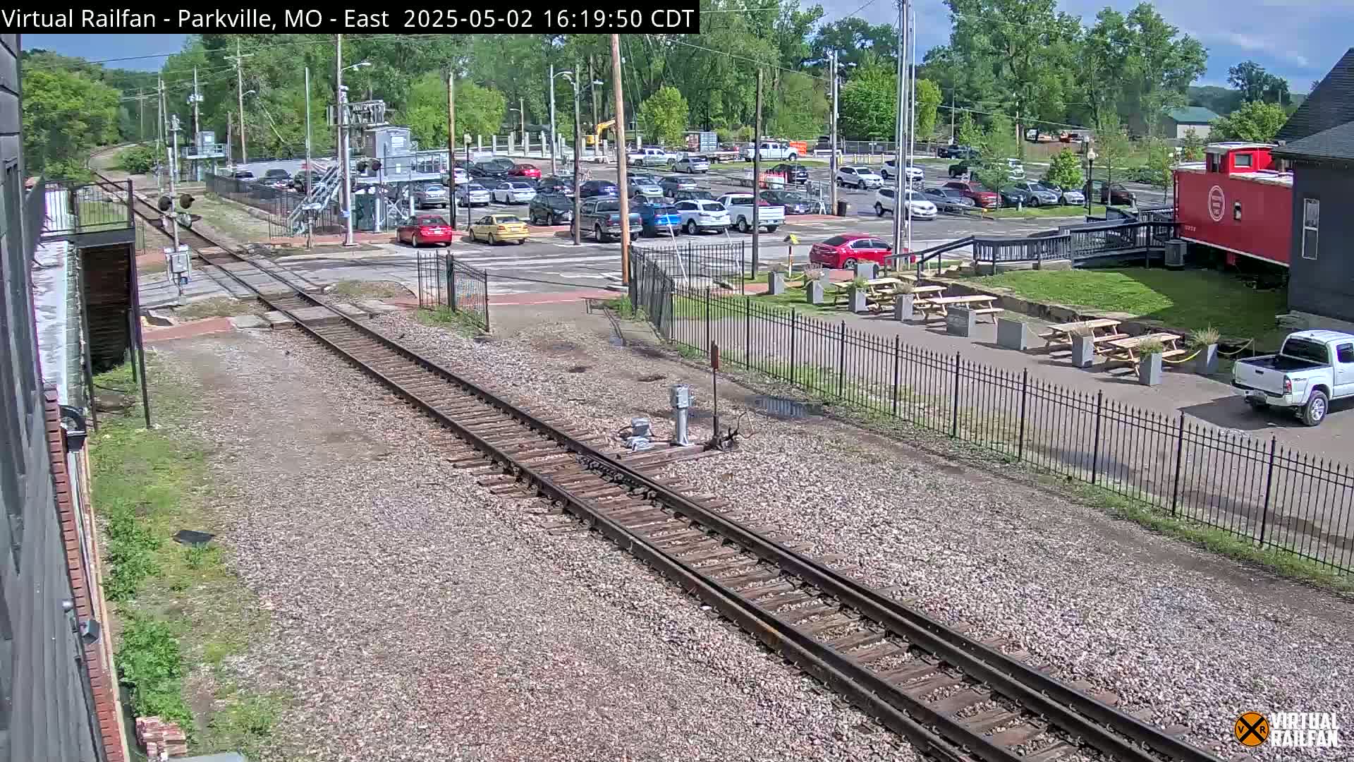 Railroad tracks intersect a street with parked cars, a nearby building, and picnic tables under a sunny sky.