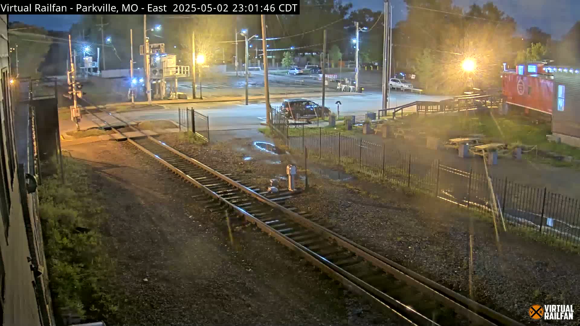 A nighttime view of railroad tracks intersecting a road, with a car parked nearby and puddles on the ground.