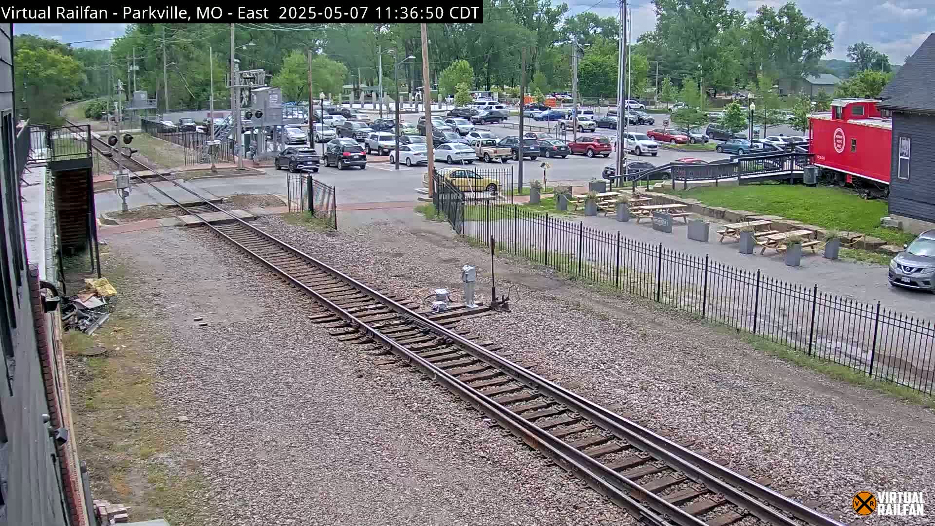 Railroad tracks pass near a parking lot, a red caboose, and several picnic tables under a partly cloudy sky.