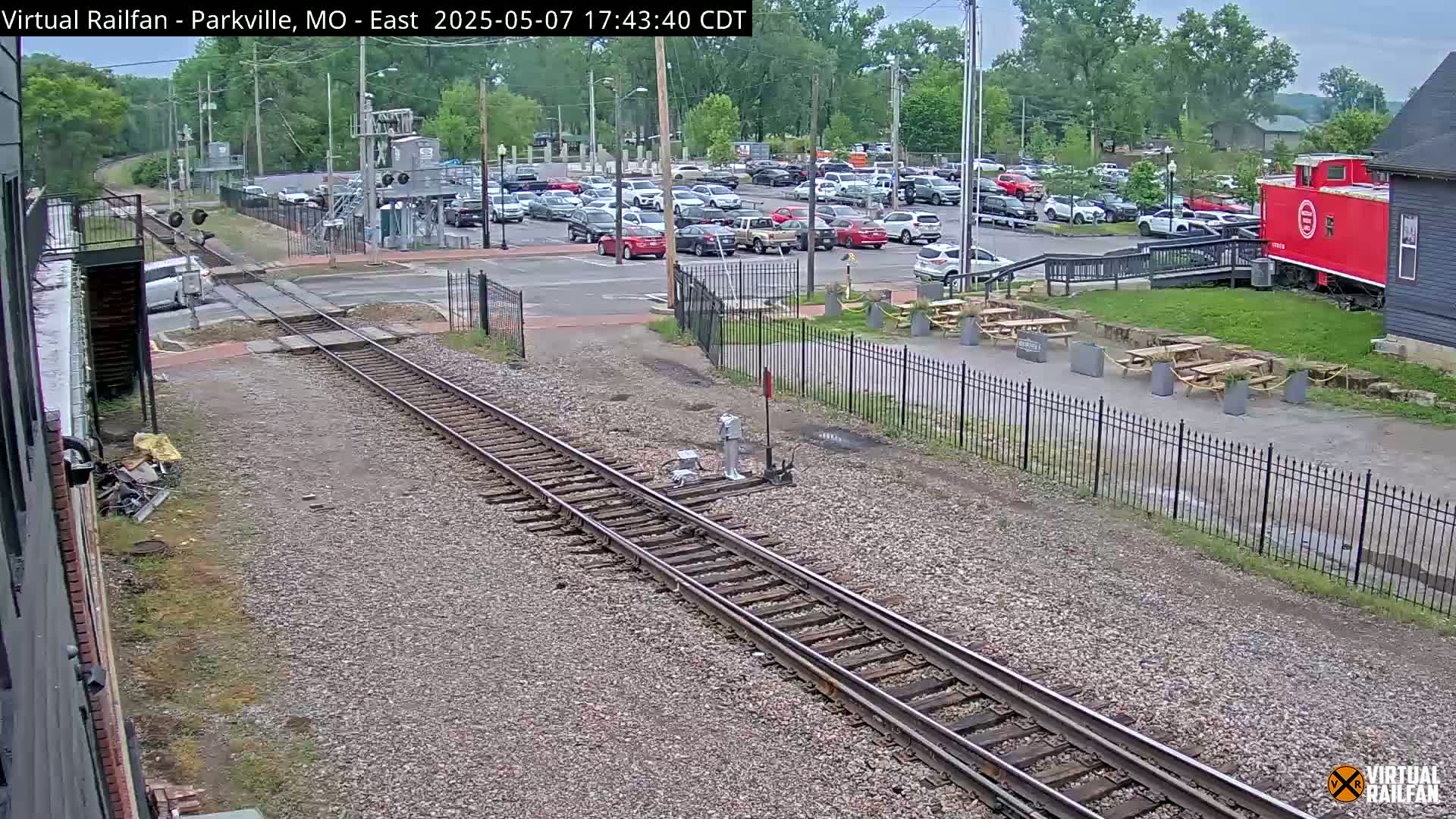 Railroad tracks extend from the foreground toward a parking lot and a red caboose next to a building, under an overcast sky.