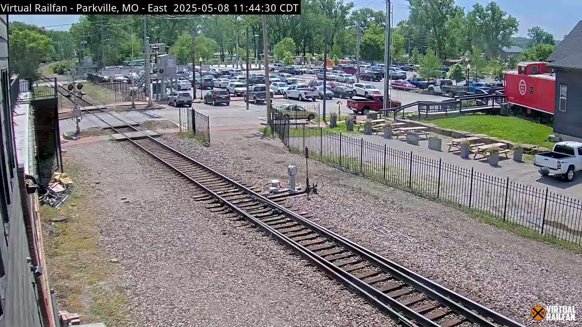 Railroad tracks cross a street next to a parking lot and outdoor seating area on a sunny day.