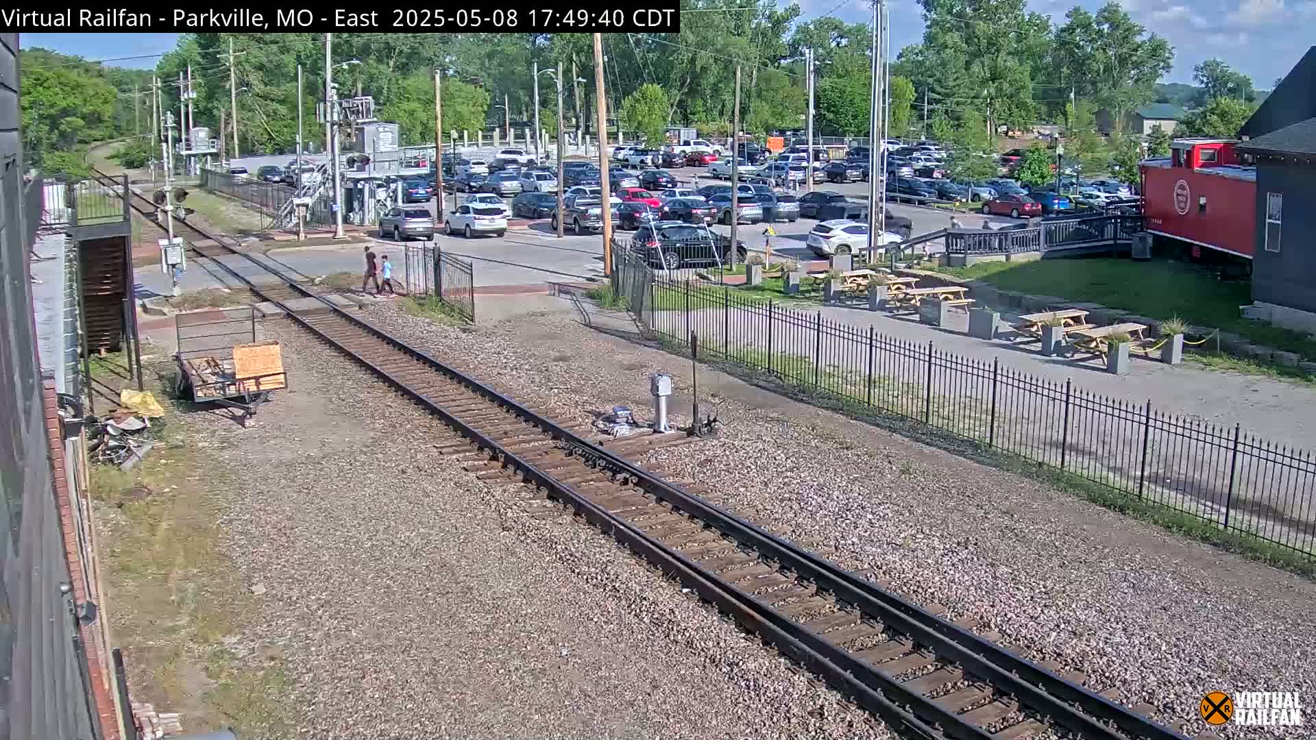 A sunny day shows railroad tracks intersecting with a parking lot full of cars near a building with outdoor seating.