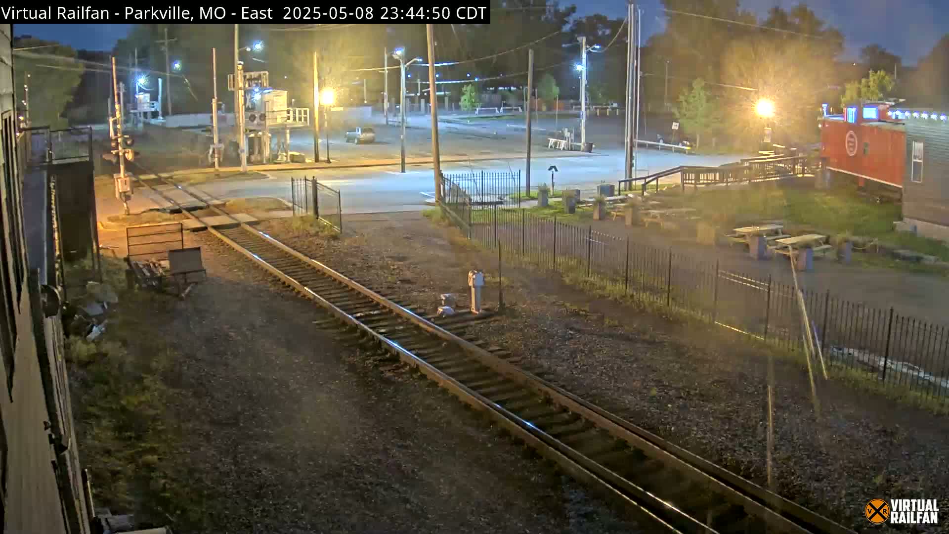 A nighttime view of railroad tracks intersecting a road near a building and picnic tables, under partly cloudy skies.