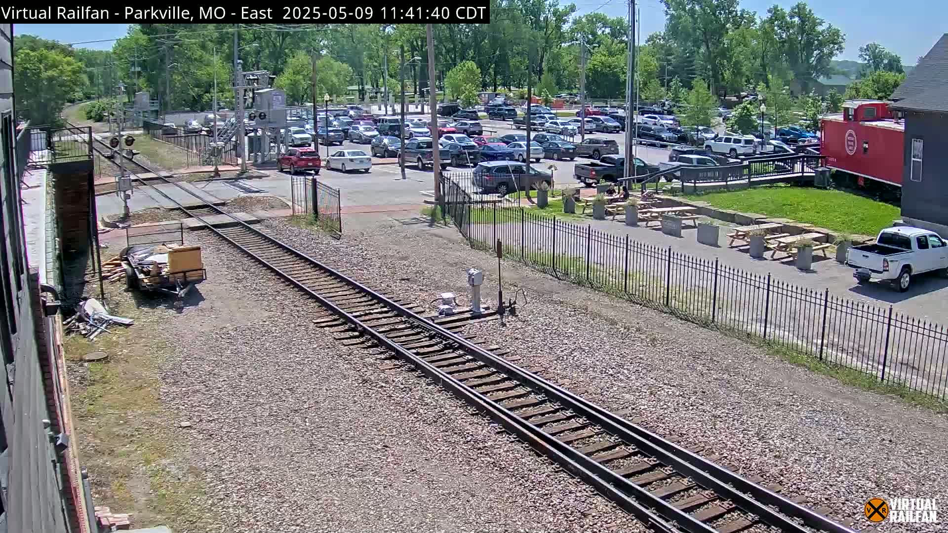 Railroad tracks run alongside a parking lot filled with cars, a red caboose, and outdoor picnic tables under a sunny sky.