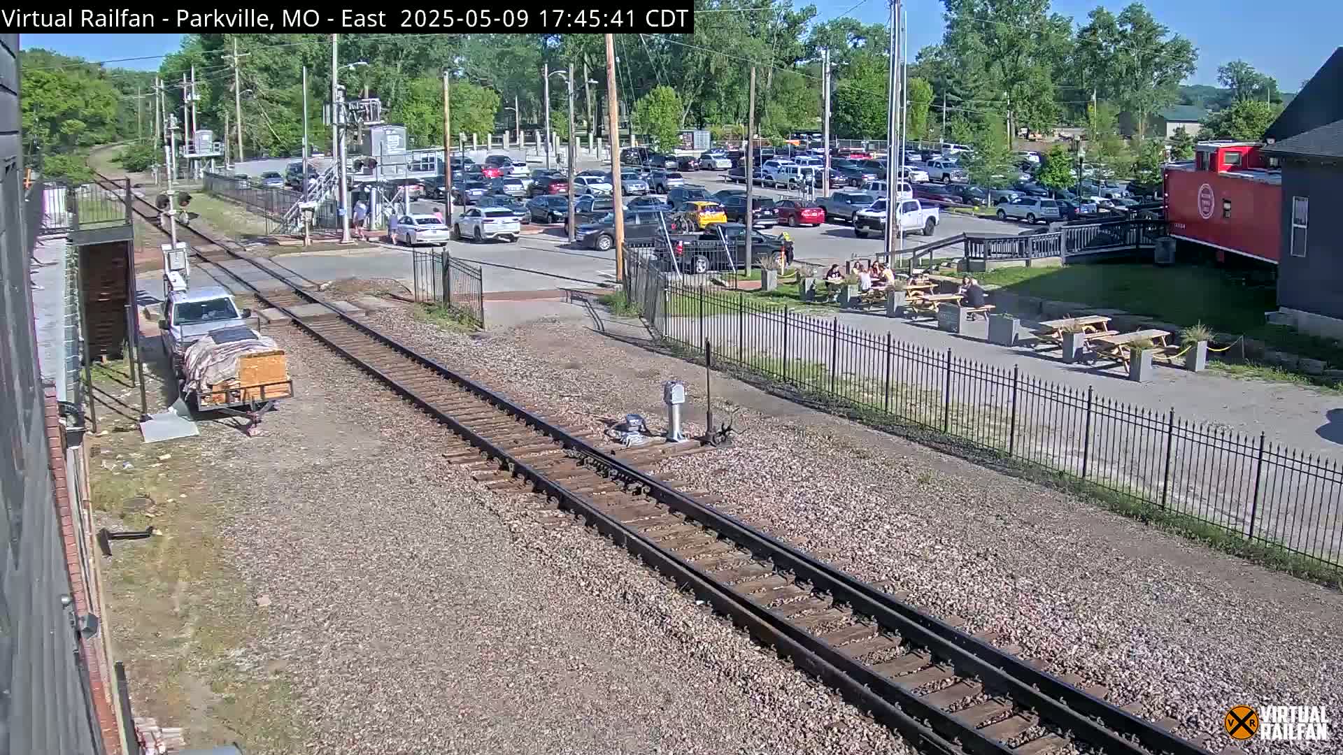 A sunny day view of railroad tracks near a parking lot and outdoor seating area, with a pickup truck and trailer parked nearby.