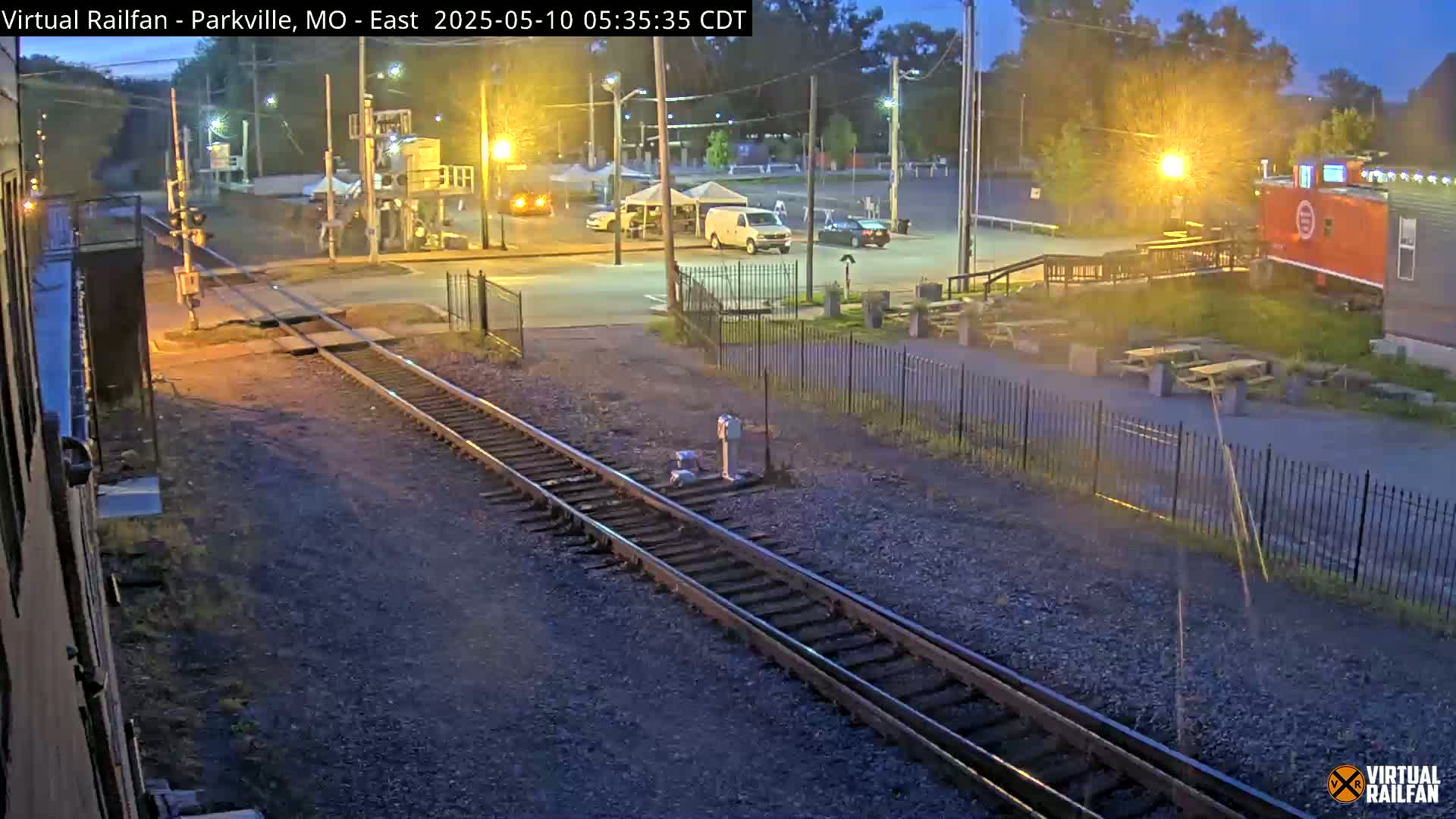 A twilight view of railroad tracks leading to a roadway with several vehicles parked nearby, and a red caboose visible in the background.