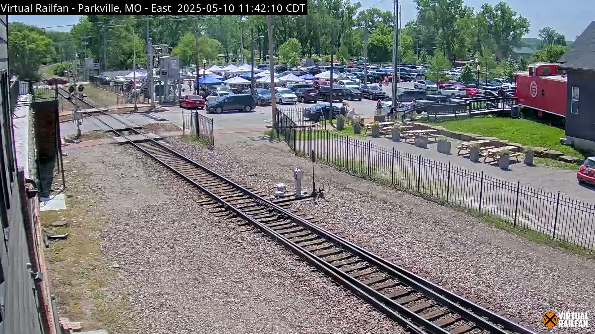 A sunny day reveals train tracks leading to a parking lot filled with cars and several white tents under a partly cloudy sky.