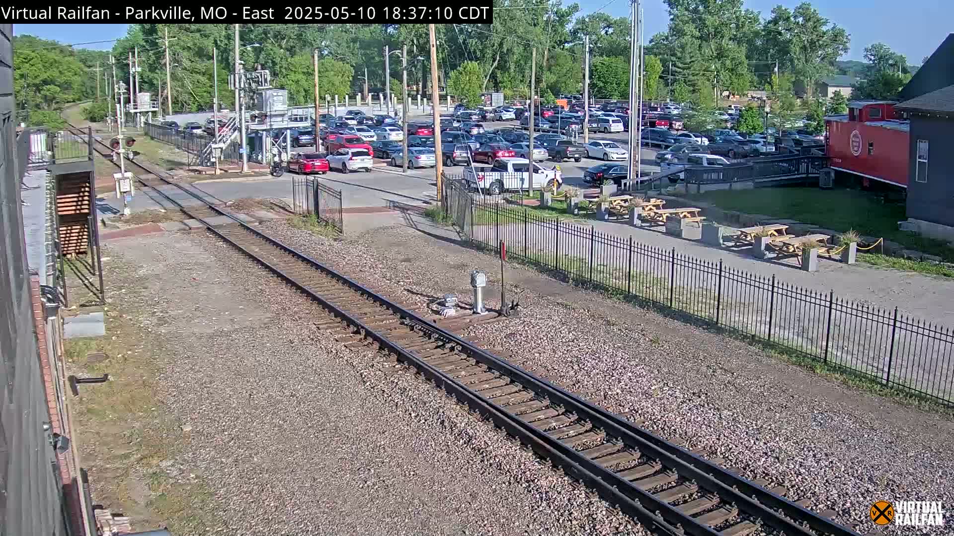 A sunny day reveals railroad tracks intersecting a roadway near a parking lot filled with cars, a red caboose, and outdoor picnic tables.