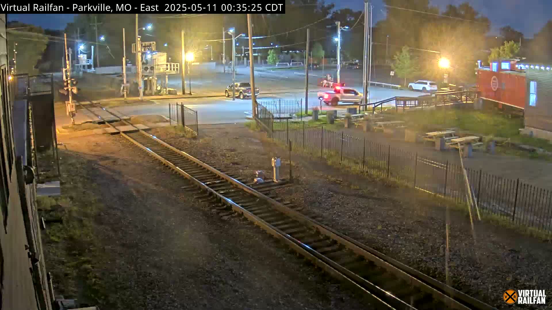 A nighttime view of railroad tracks intersecting a road with a few parked cars and a red boxcar visible in the background under clear skies.