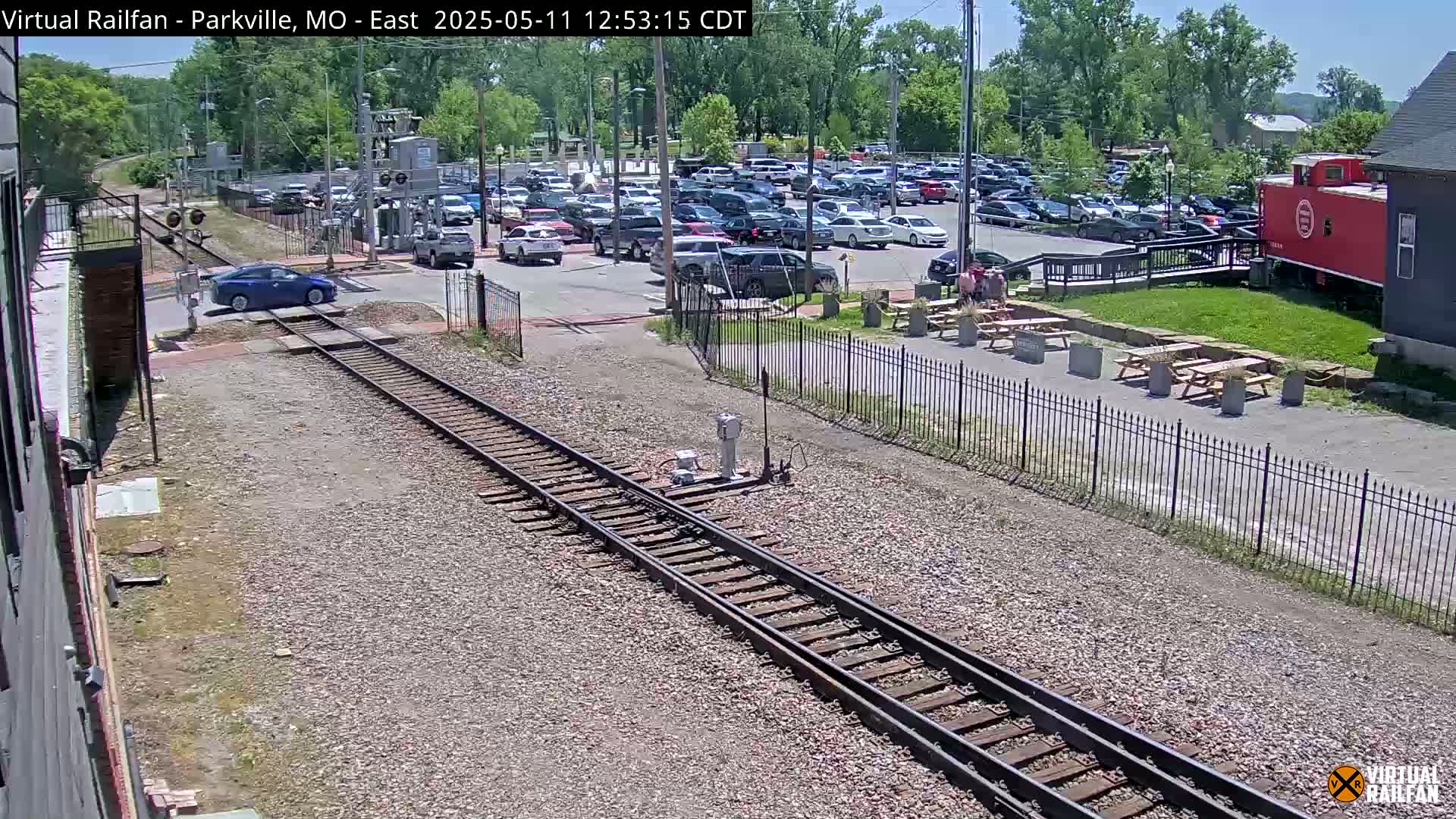 A blue car crosses a railroad track in a sunny outdoor setting near a parking lot, picnic tables, and a red caboose.