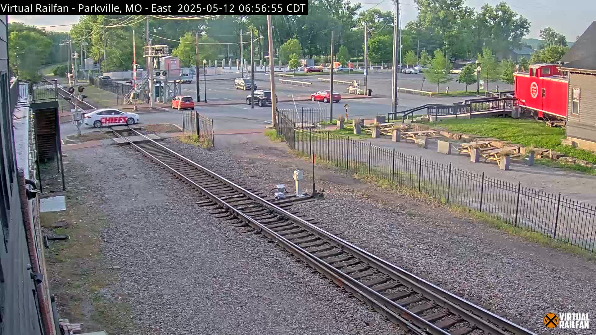 A white car with writing on its side is crossing railroad tracks in a parking lot next to a red caboose on a sunny day.