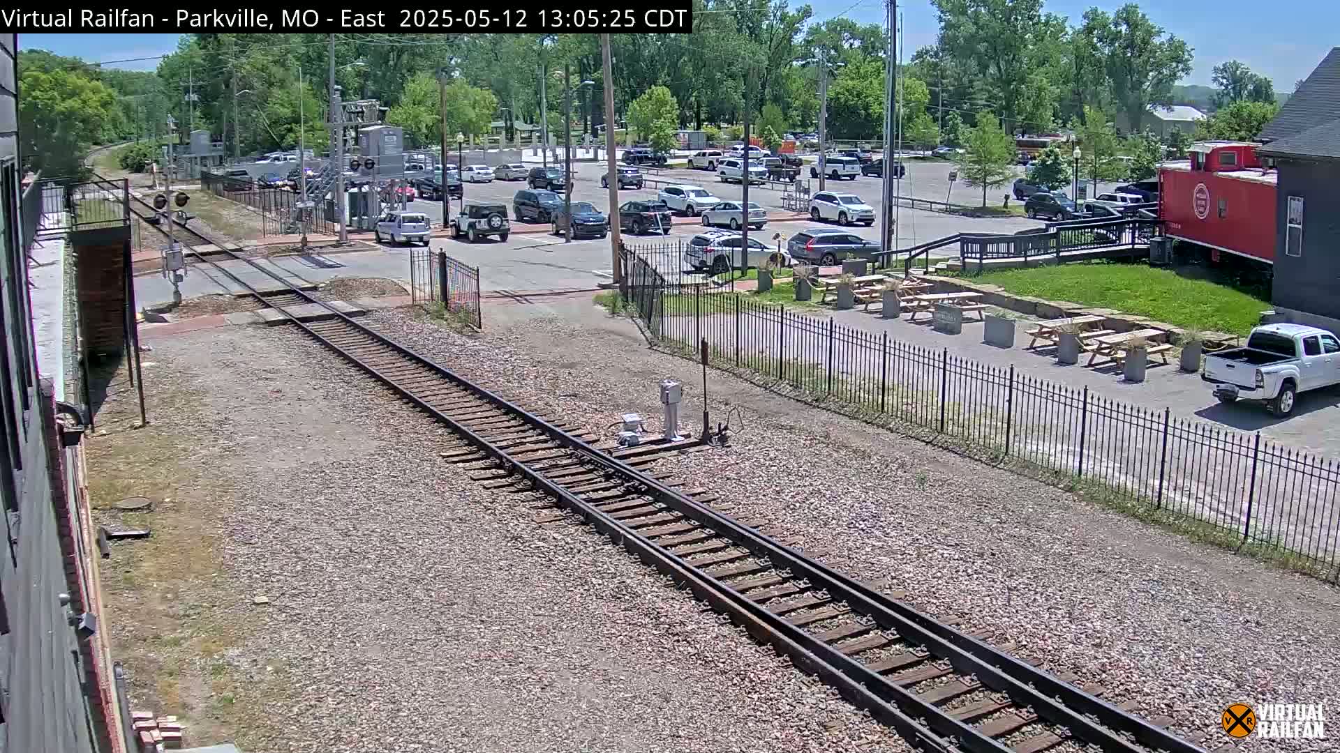 A sunny day view of railroad tracks, a parking lot filled with cars, and a red caboose near picnic tables and a black building.