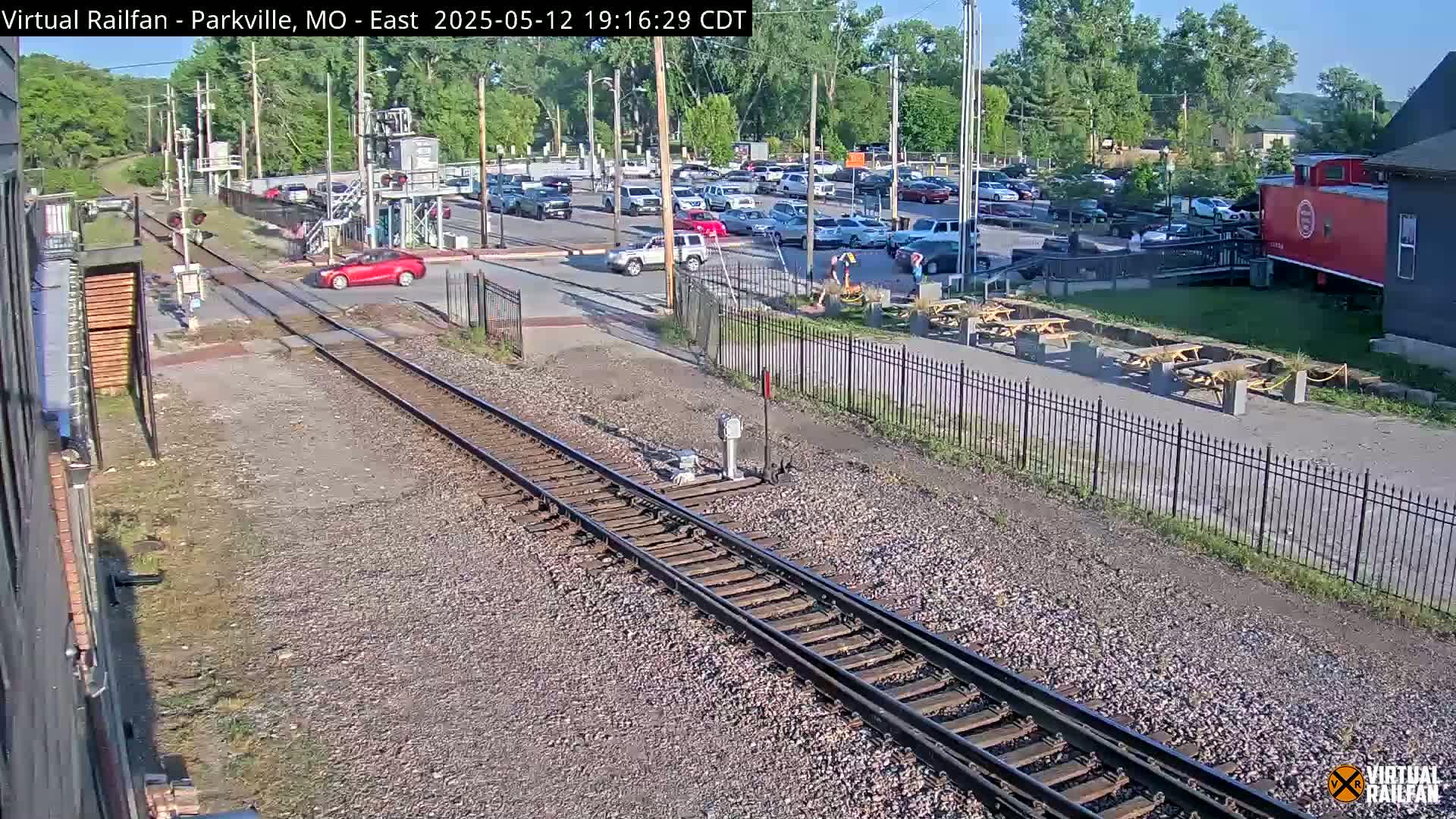 A sunny day view of railroad tracks crossing a road near a parking lot with several cars and a red caboose, next to outdoor seating and a building.