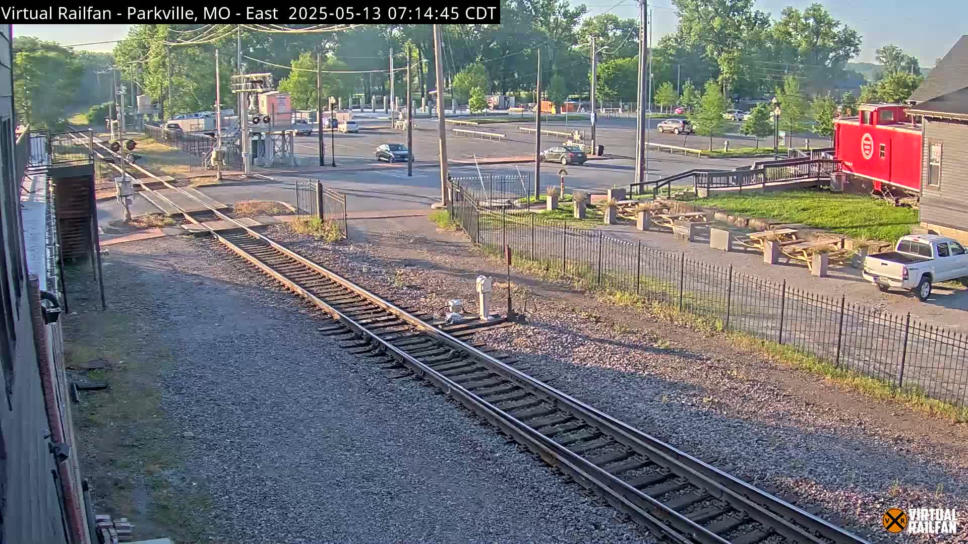 A sunny view of railroad tracks intersecting with a roadway, near a red caboose and a parking lot with several cars.