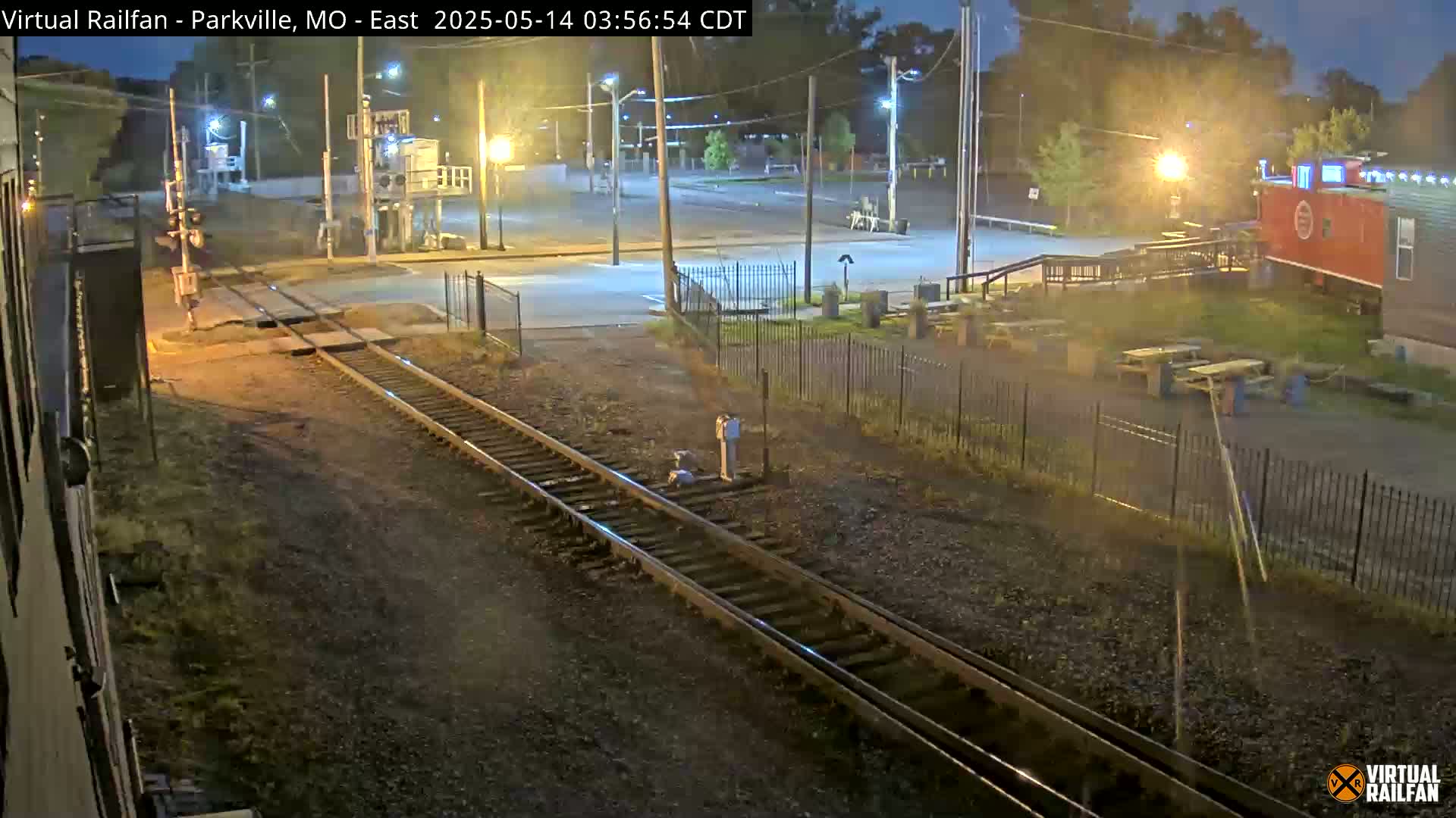 At night, railroad tracks cross a street near a fenced-in area with picnic tables and a red building.