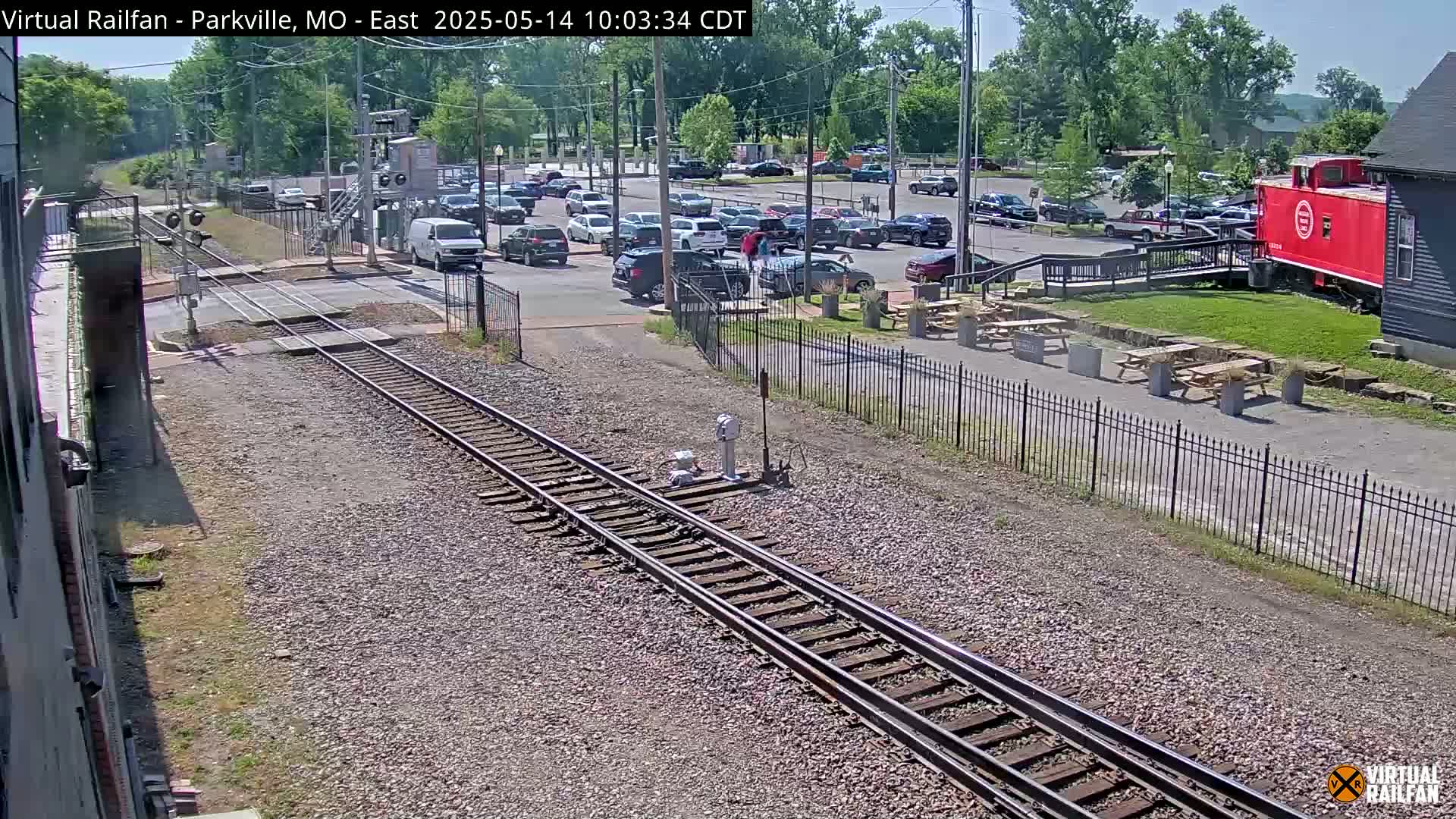 A sunny day shows a railroad crossing with a parking lot full of cars and a red caboose next to a building.