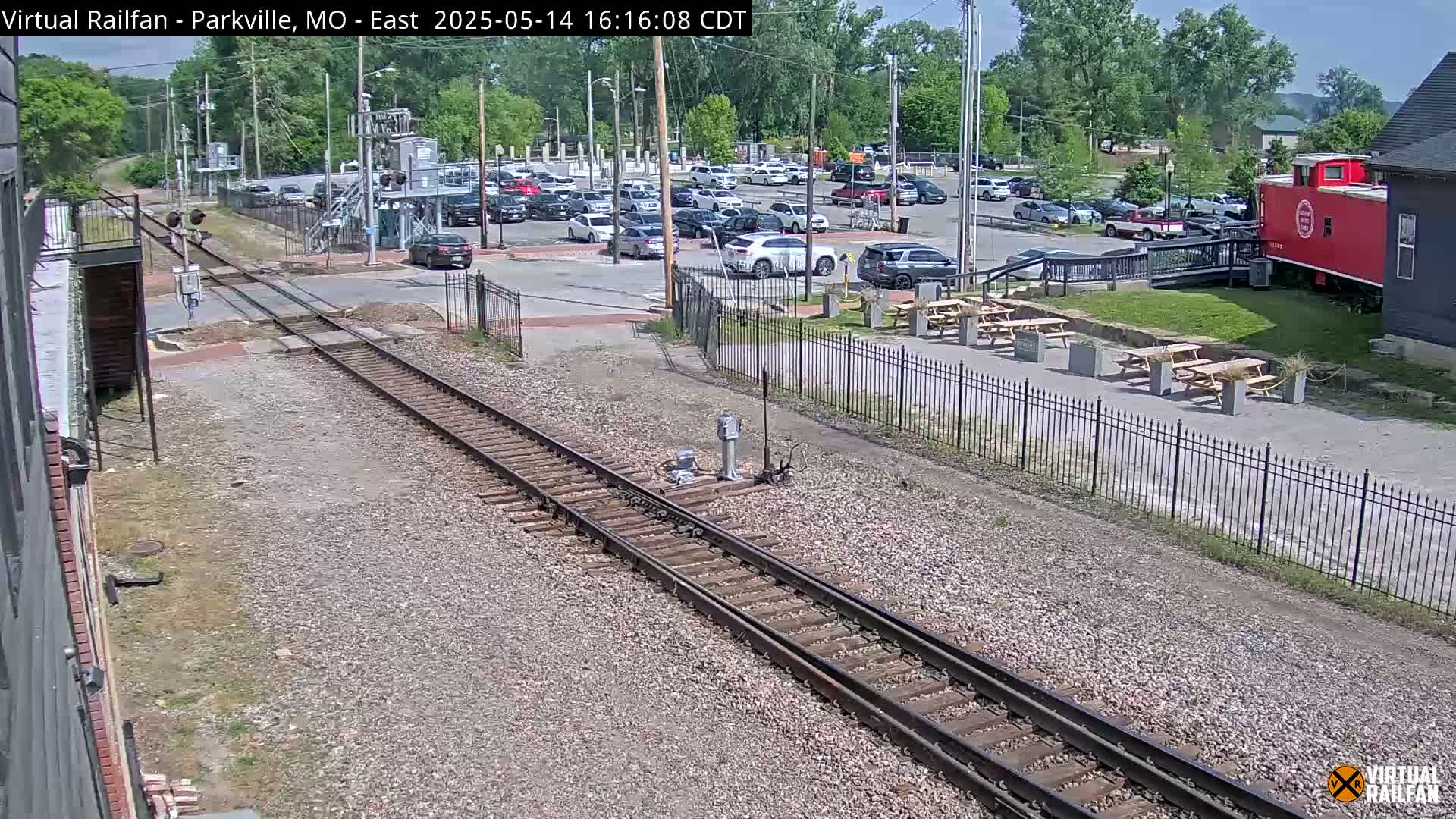 Railroad tracks run through a parking lot with cars parked, picnic tables outside a building, and a red caboose visible in the background on a sunny day.