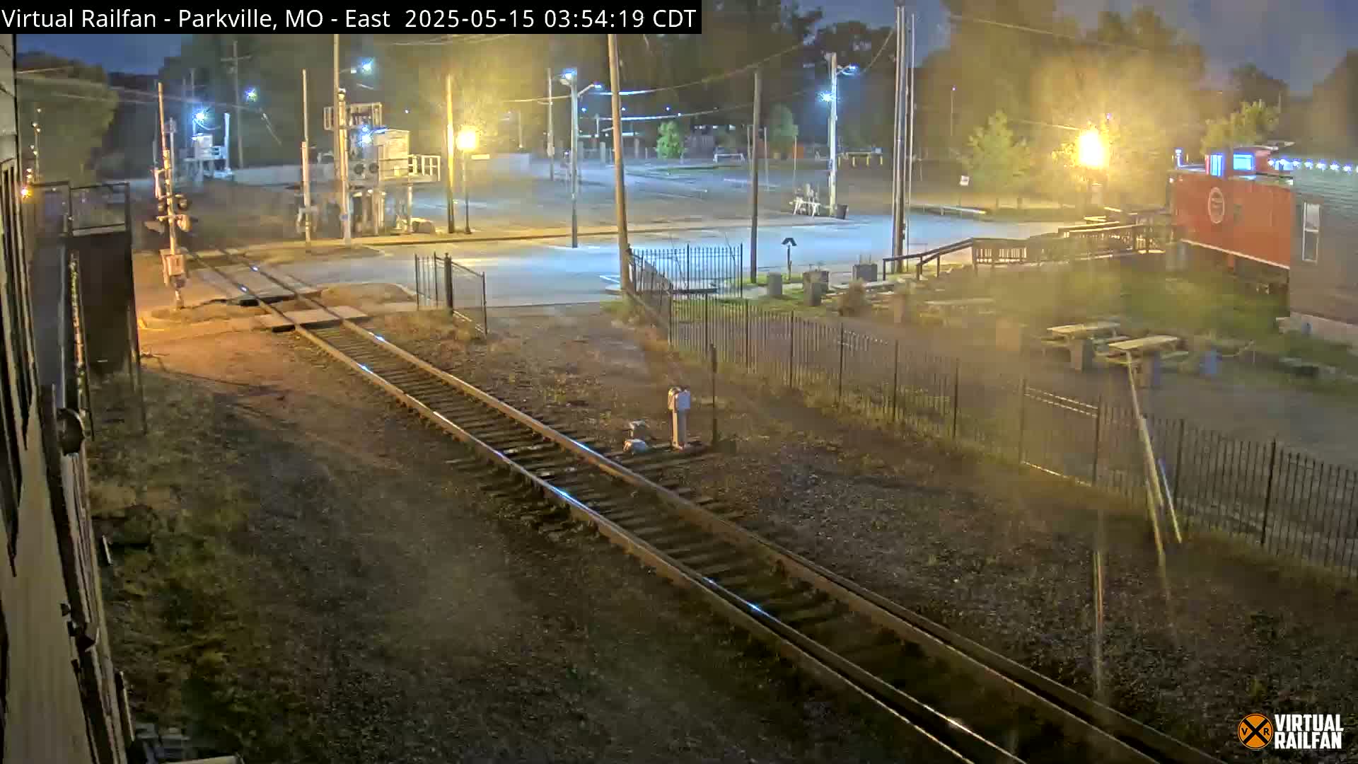 A nighttime view of railroad tracks intersecting a road, with a red caboose visible in the background under dim streetlights.