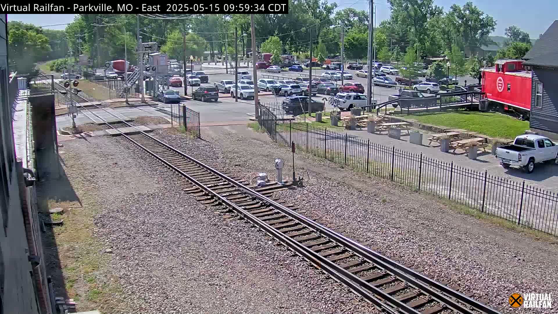 A sunny day shows train tracks intersecting a road with parked cars and a red caboose visible near a building and picnic tables.