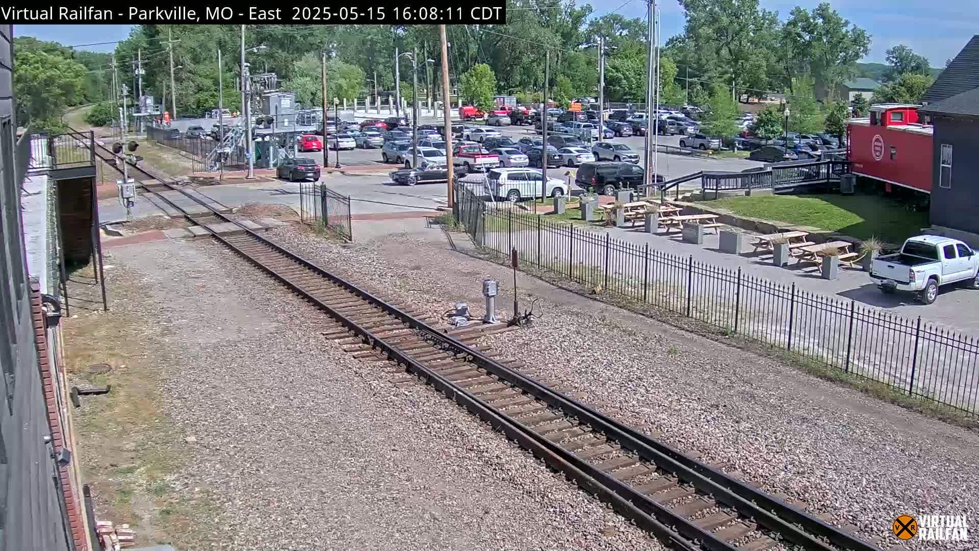 A sunny day shows a railroad crossing near a parking lot with several cars and picnic tables next to a red caboose.