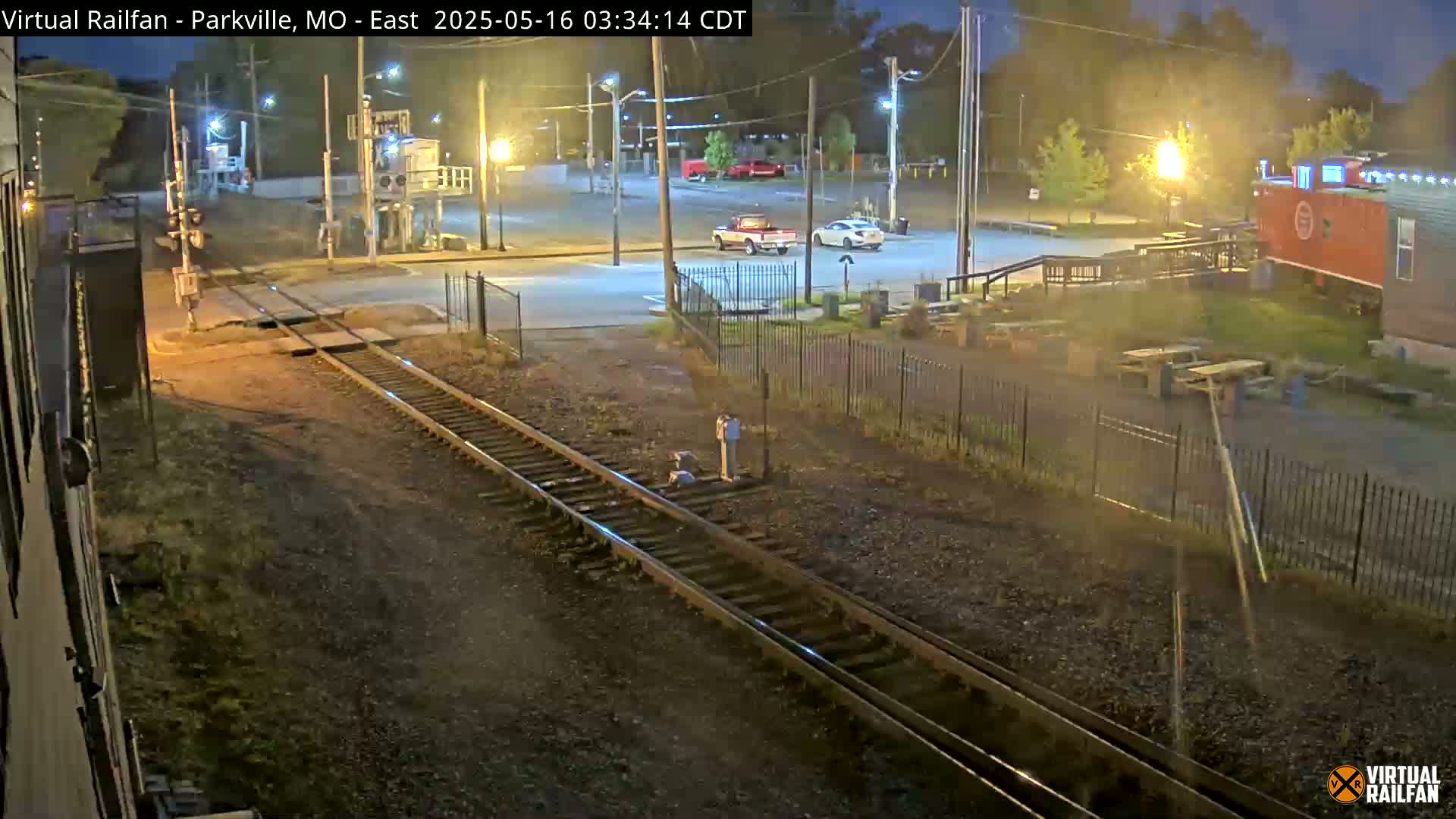 A nighttime view of railroad tracks intersecting a road, with a few vehicles parked in a lot nearby, and a red caboose-like structure visible in the background.