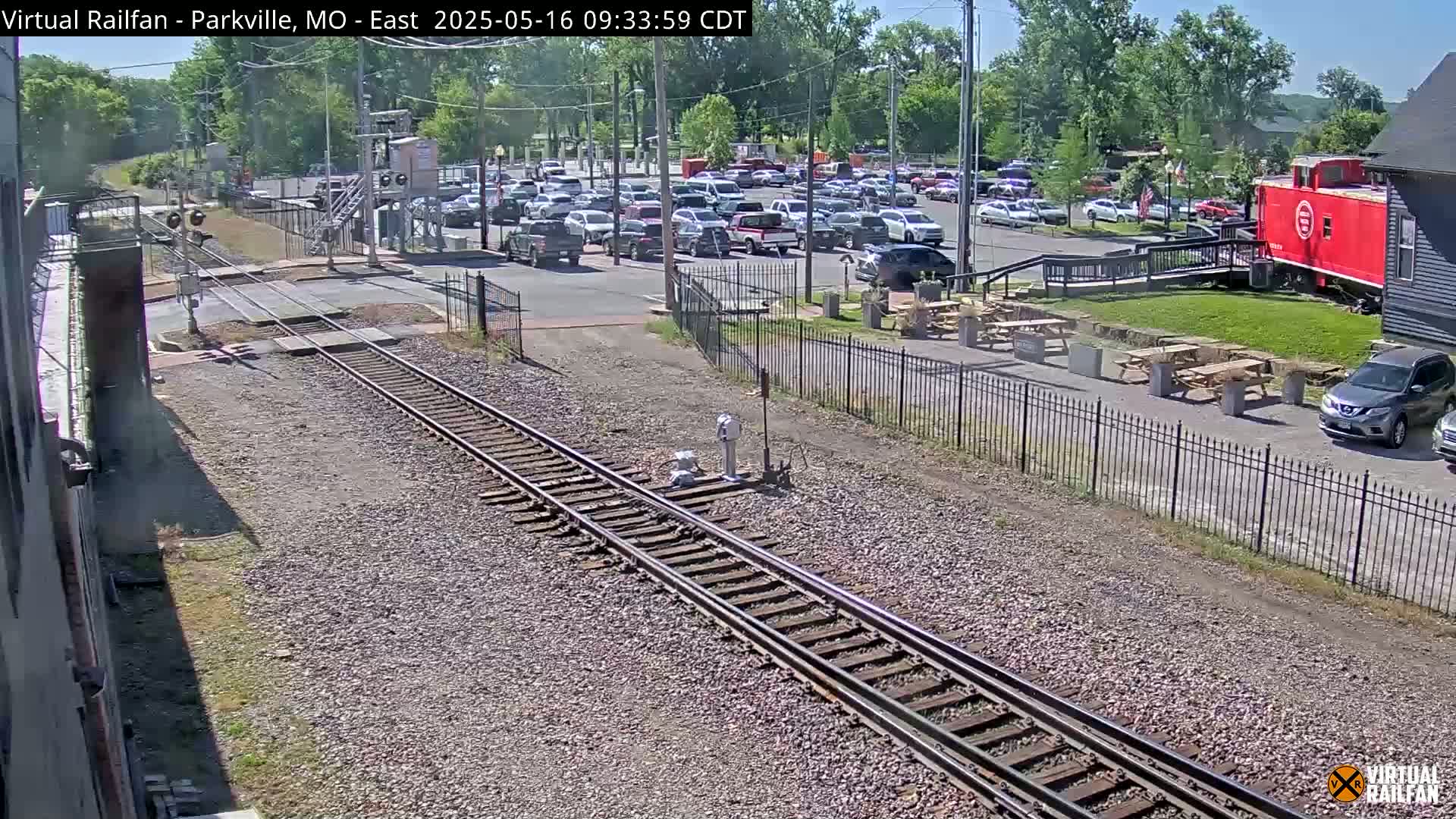 A sunny outdoor view shows railroad tracks leading to a parking lot filled with cars, a red caboose, and several picnic tables.