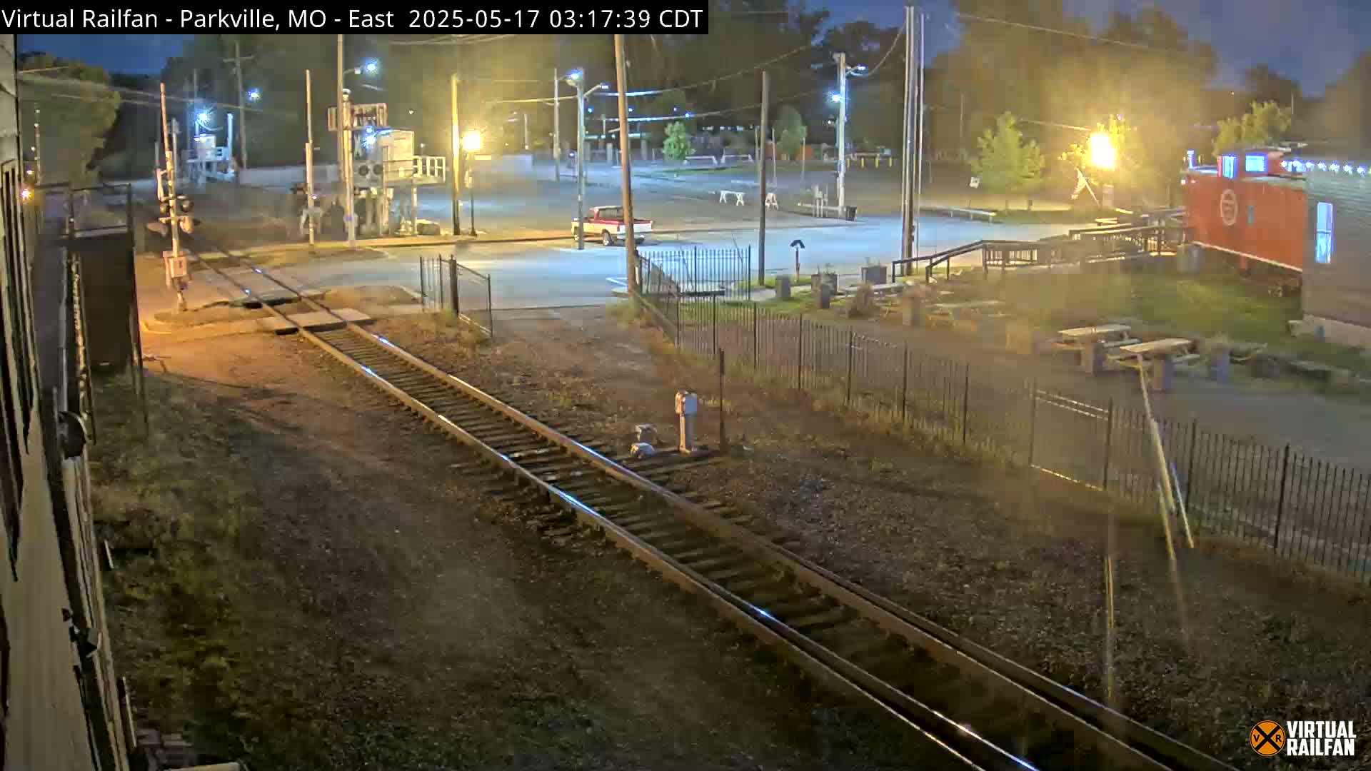 A nighttime view of railroad tracks intersecting a road, with a pickup truck parked nearby, and a red caboose visible in the background under streetlights.