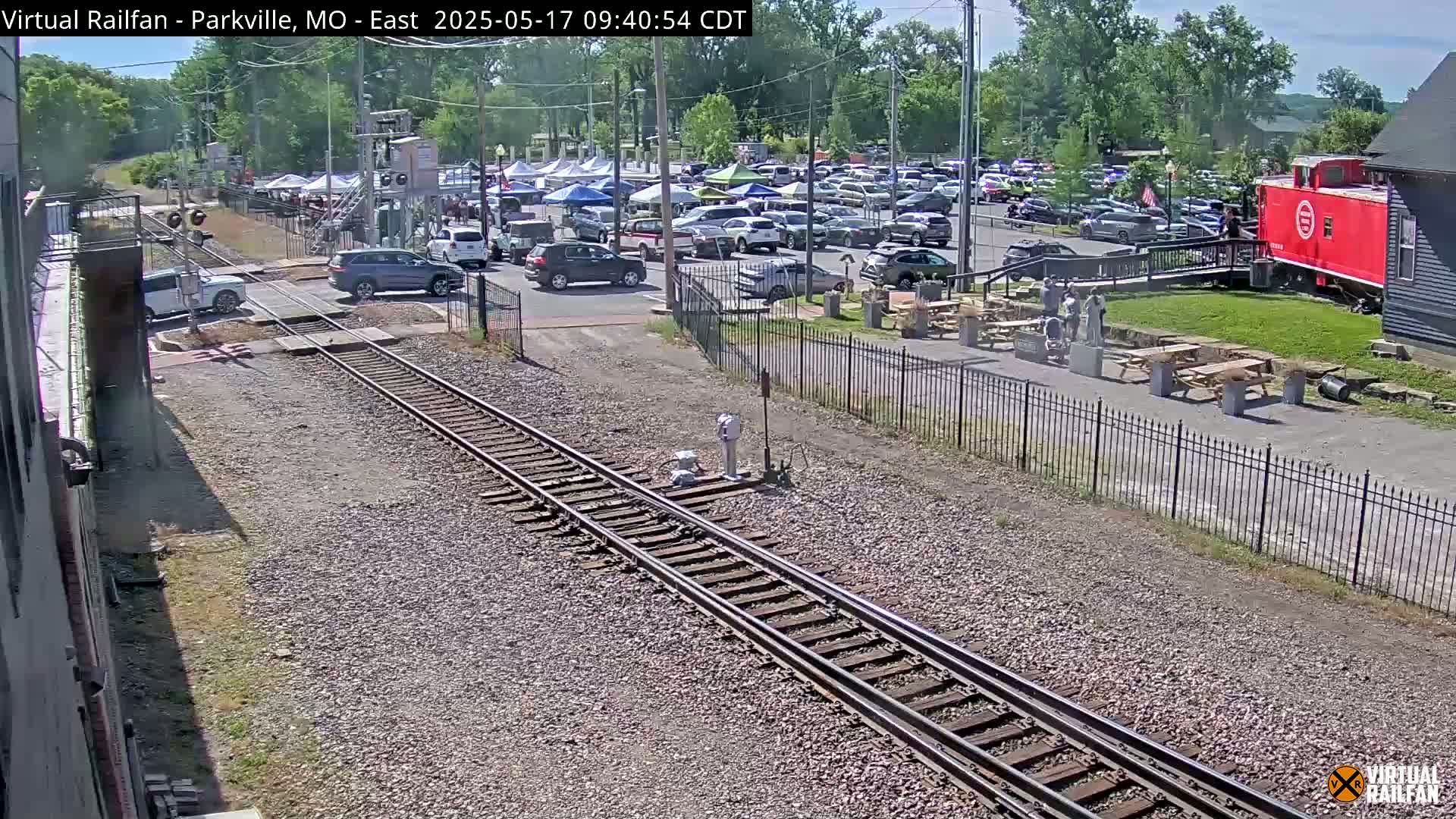 A sunny day shows railroad tracks intersecting with a road where several cars are parked near a red caboose and picnic tables.