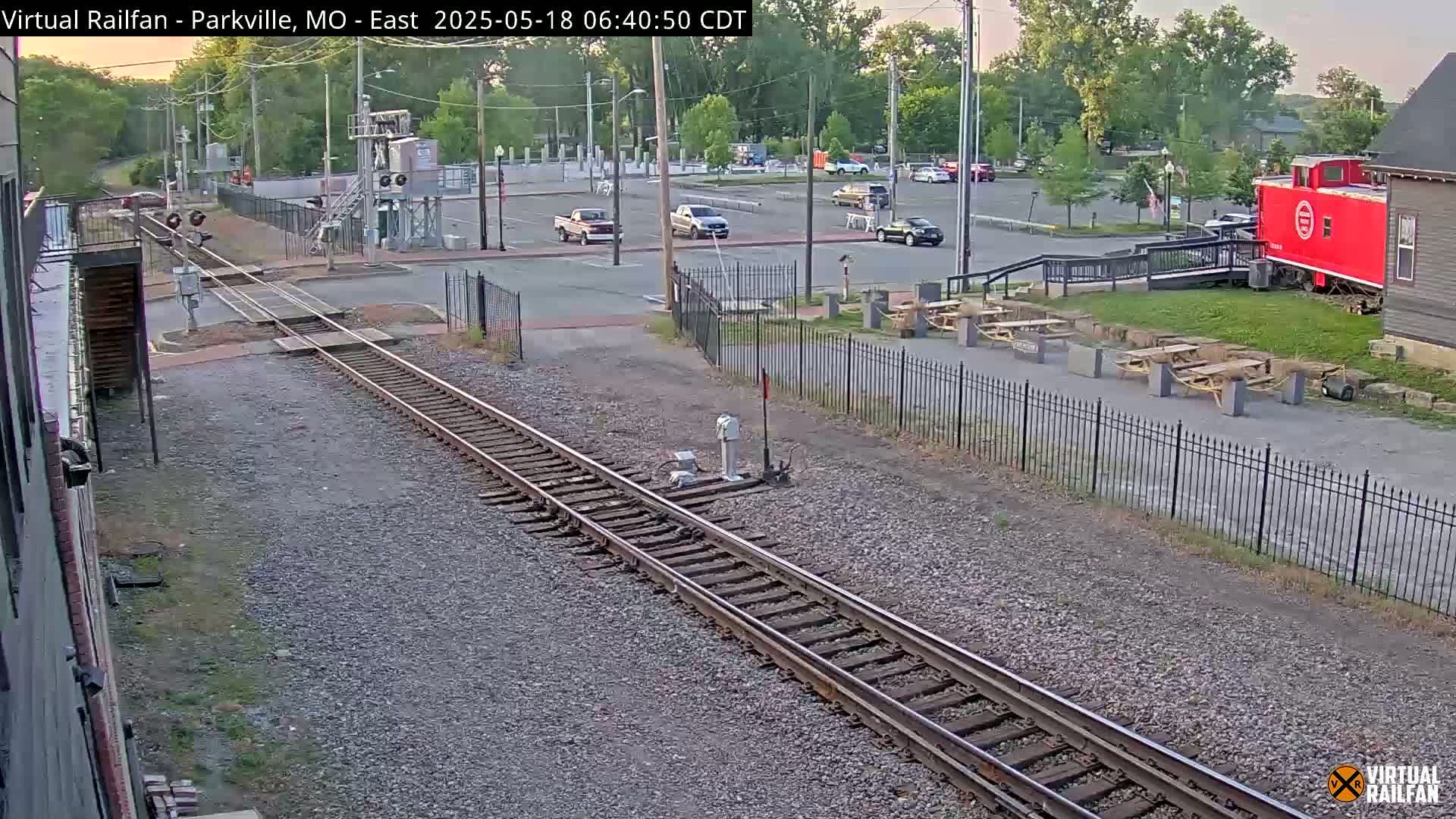 Railroad tracks extend from the foreground toward a parking lot with several cars, a red caboose, and picnic tables under a partly sunny sky.