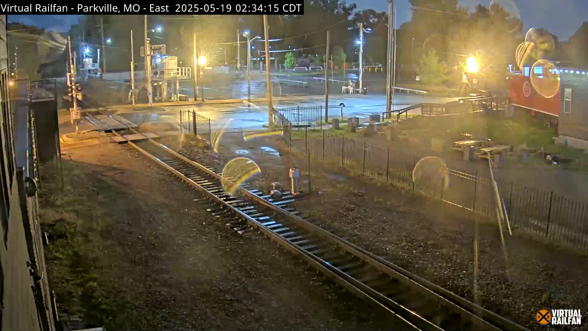 A nighttime view of a railway crossing and adjacent area, showing wet pavement under light rain.