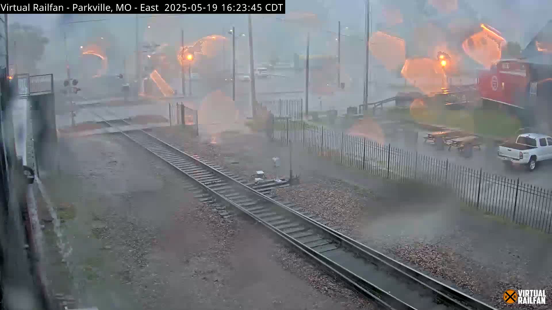 A view of railroad tracks during a heavy rainstorm shows a red train car in the distance and a white truck parked nearby.