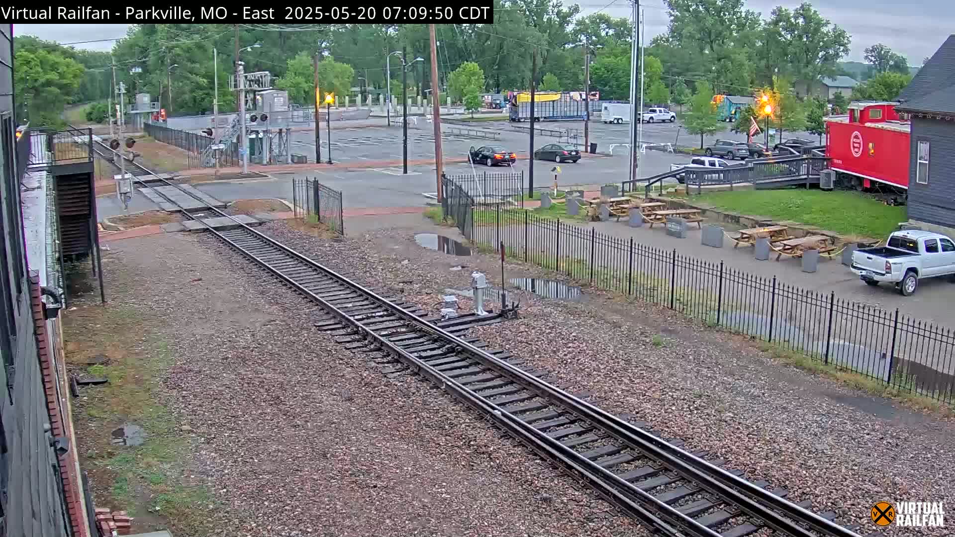 A slightly overcast view of railroad tracks next to a parking lot with several cars and picnic tables, near a red boxcar and a building.