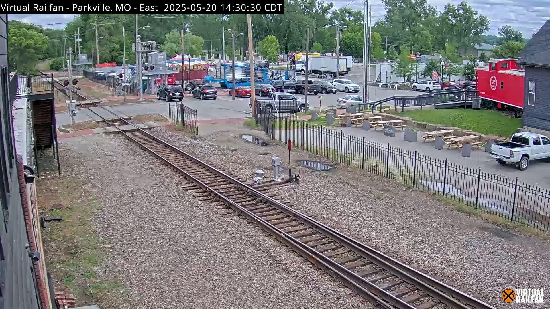 A railroad track runs through a parking lot with several cars and a red caboose next to picnic tables under a partly cloudy sky.