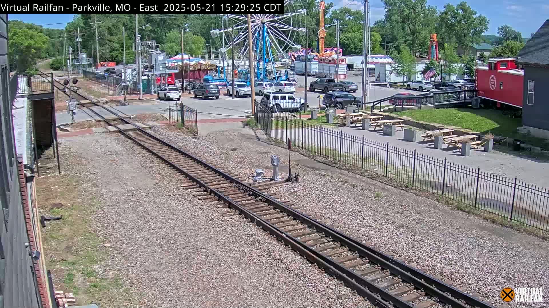 Railroad tracks run alongside a parking lot with several cars and a Ferris wheel under a sunny sky.