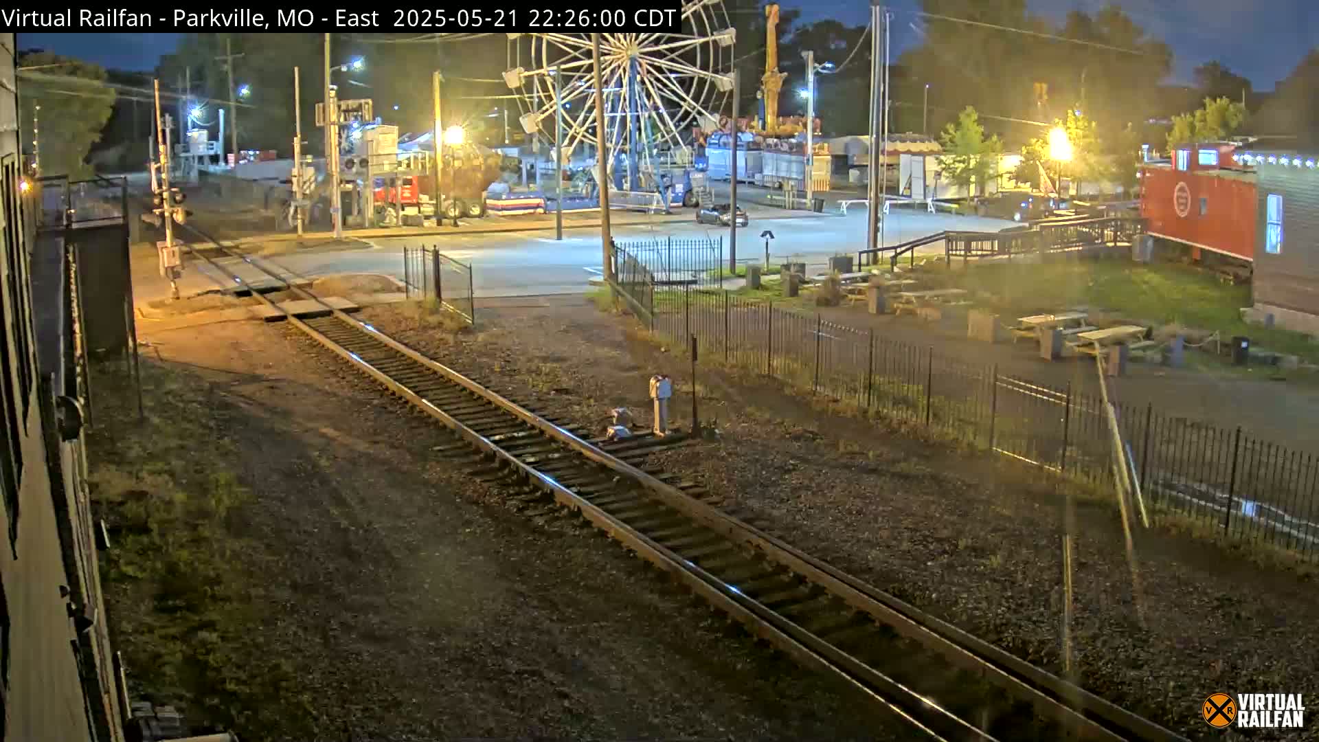 A nighttime view of railroad tracks leading towards a Ferris wheel and carnival rides, with a red caboose visible in the background.