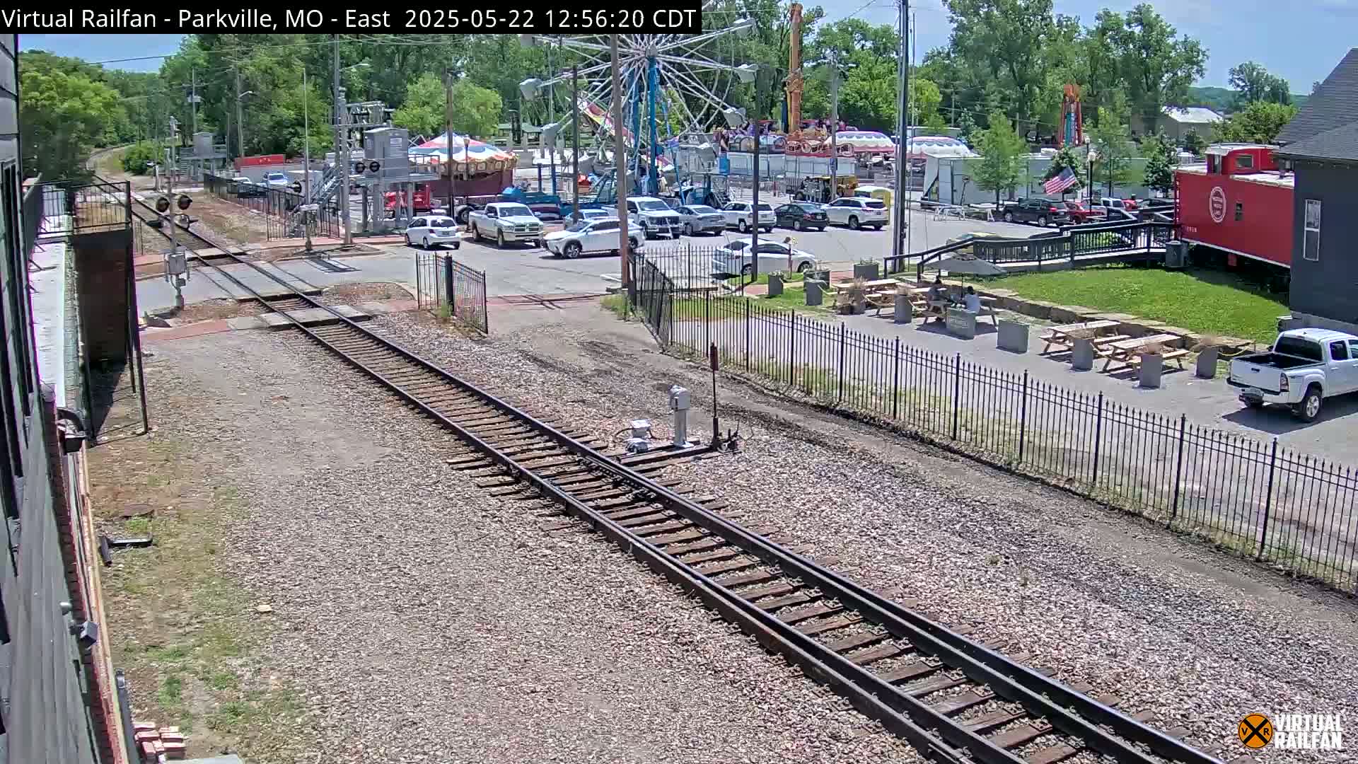 Railroad tracks run through a sunny area with a carnival, parked cars, and picnic tables near a red caboose.