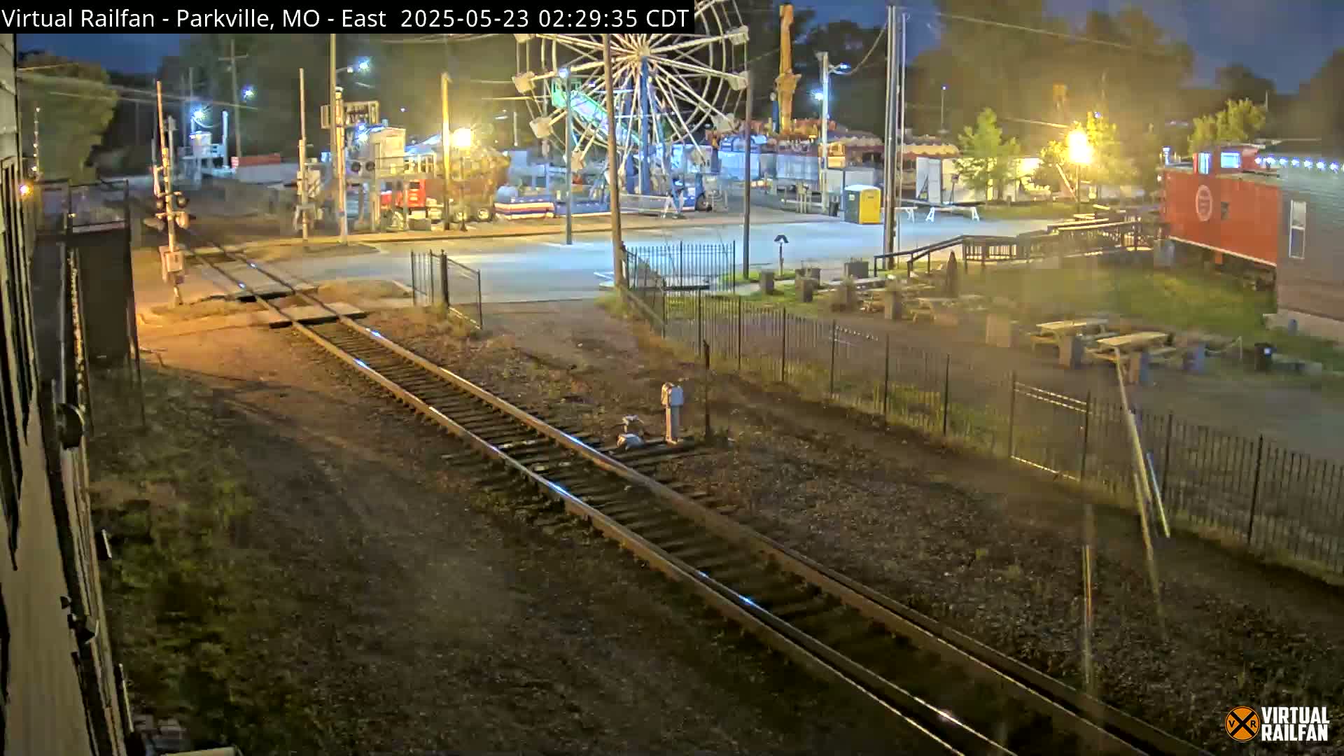 At night, a view from a train shows railroad tracks leading toward a carnival-like area with a Ferris wheel, some buildings, and picnic tables.