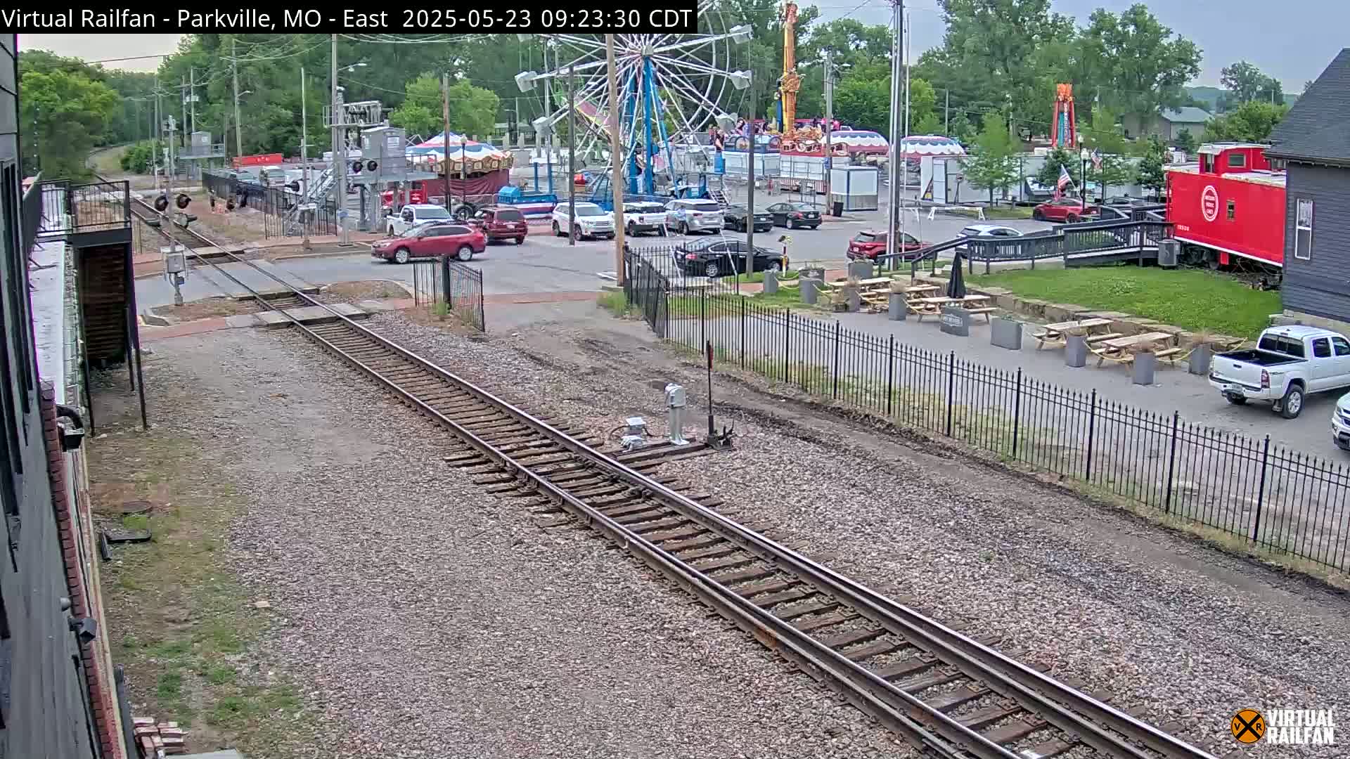 A view of railroad tracks intersecting with a street, showing a carnival in the background on an overcast day.