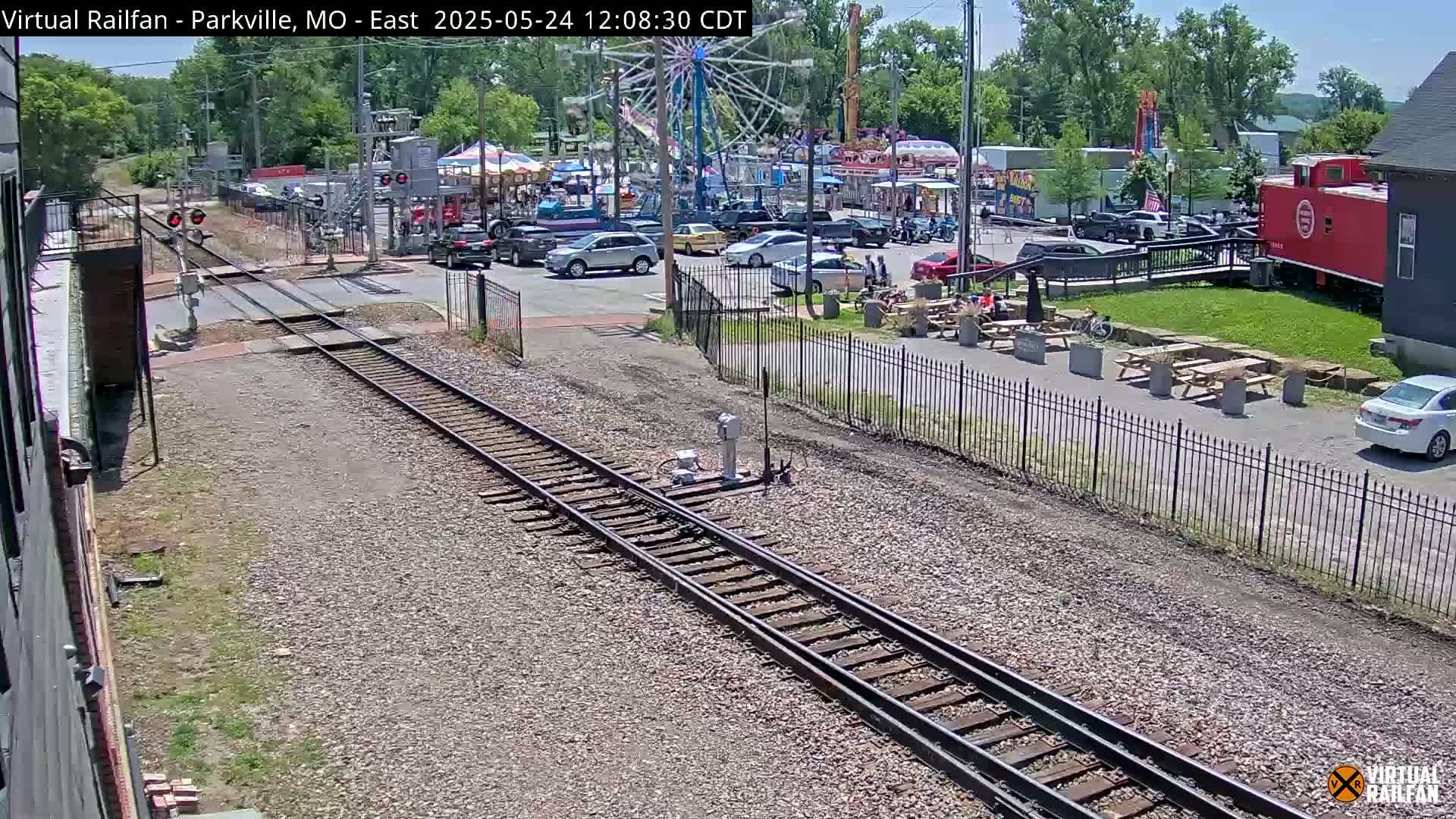 A sunny day shows railroad tracks intersecting a road next to a fairground with parked cars, picnic tables, and a red caboose.