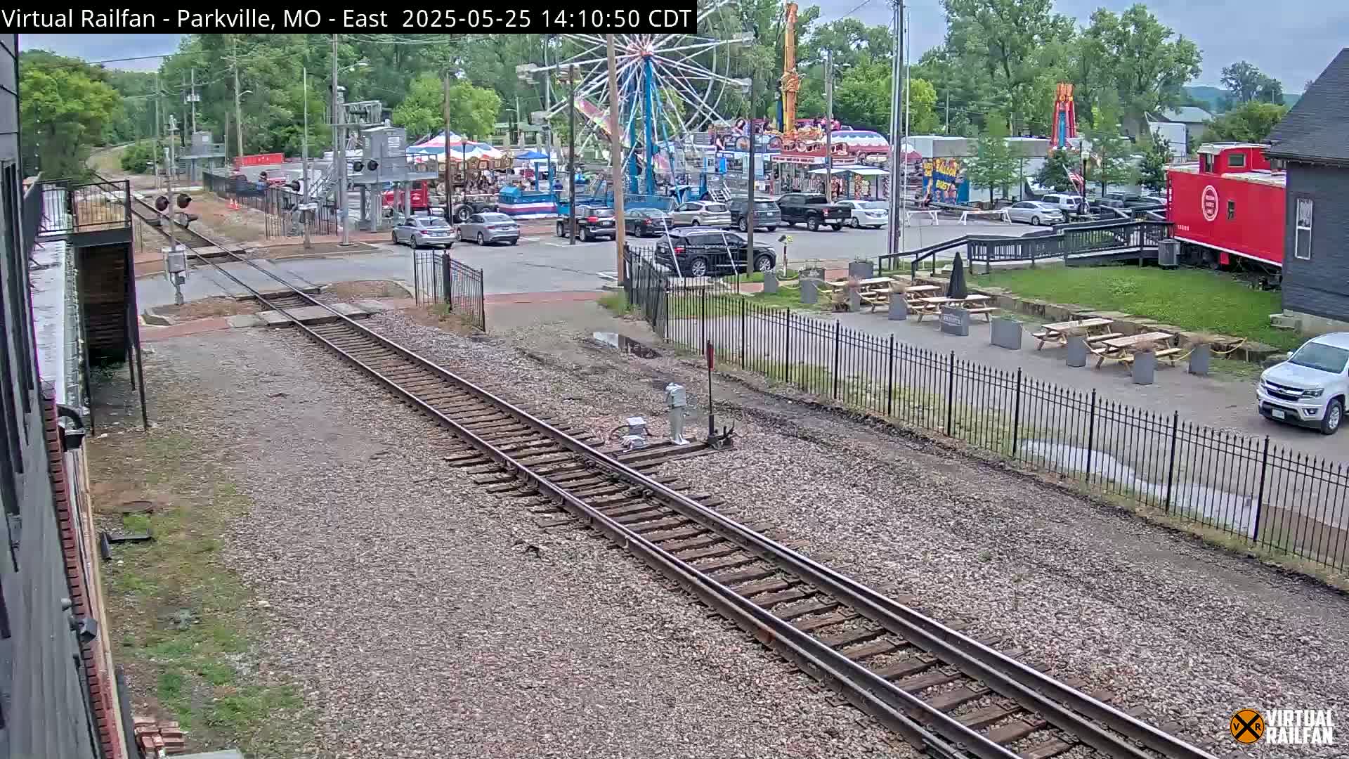 Train tracks run alongside a street with parked cars near a carnival, under a partly cloudy sky.