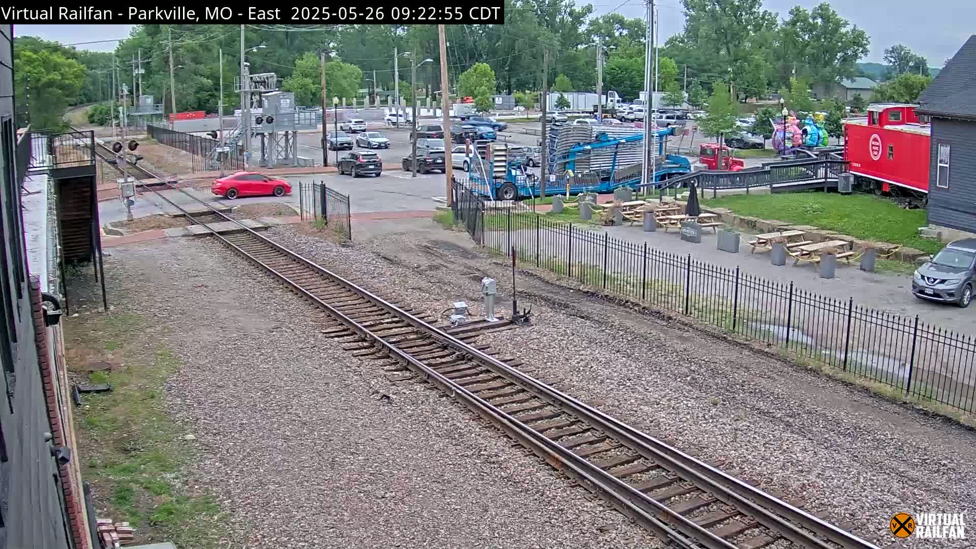 A red car crosses railroad tracks near a red caboose and several picnic tables, under an overcast sky.